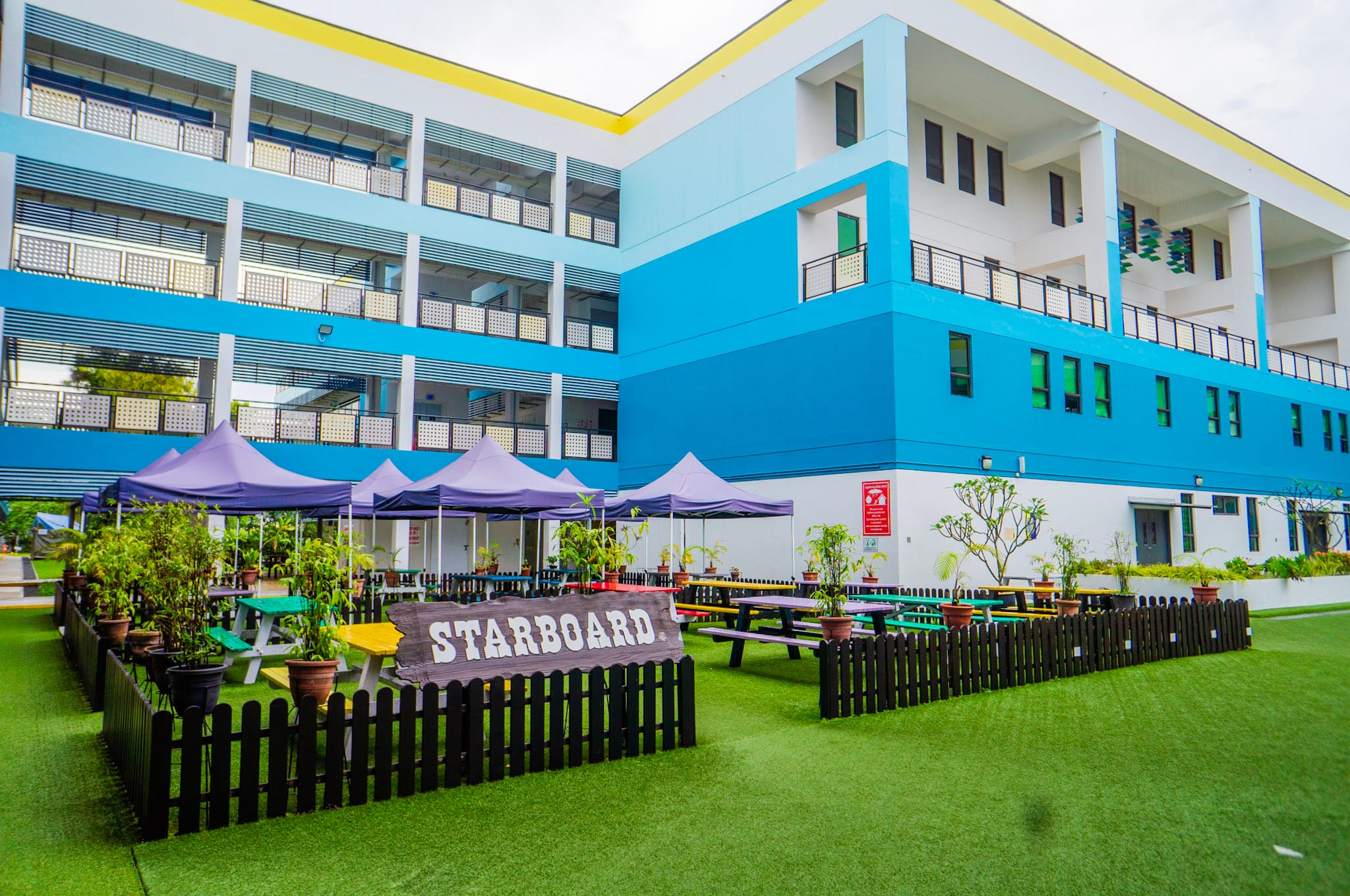 Blue multi-story school building with a fenced outdoor area labeled Starboard, colorful benches, purple canopies, and potted plants.