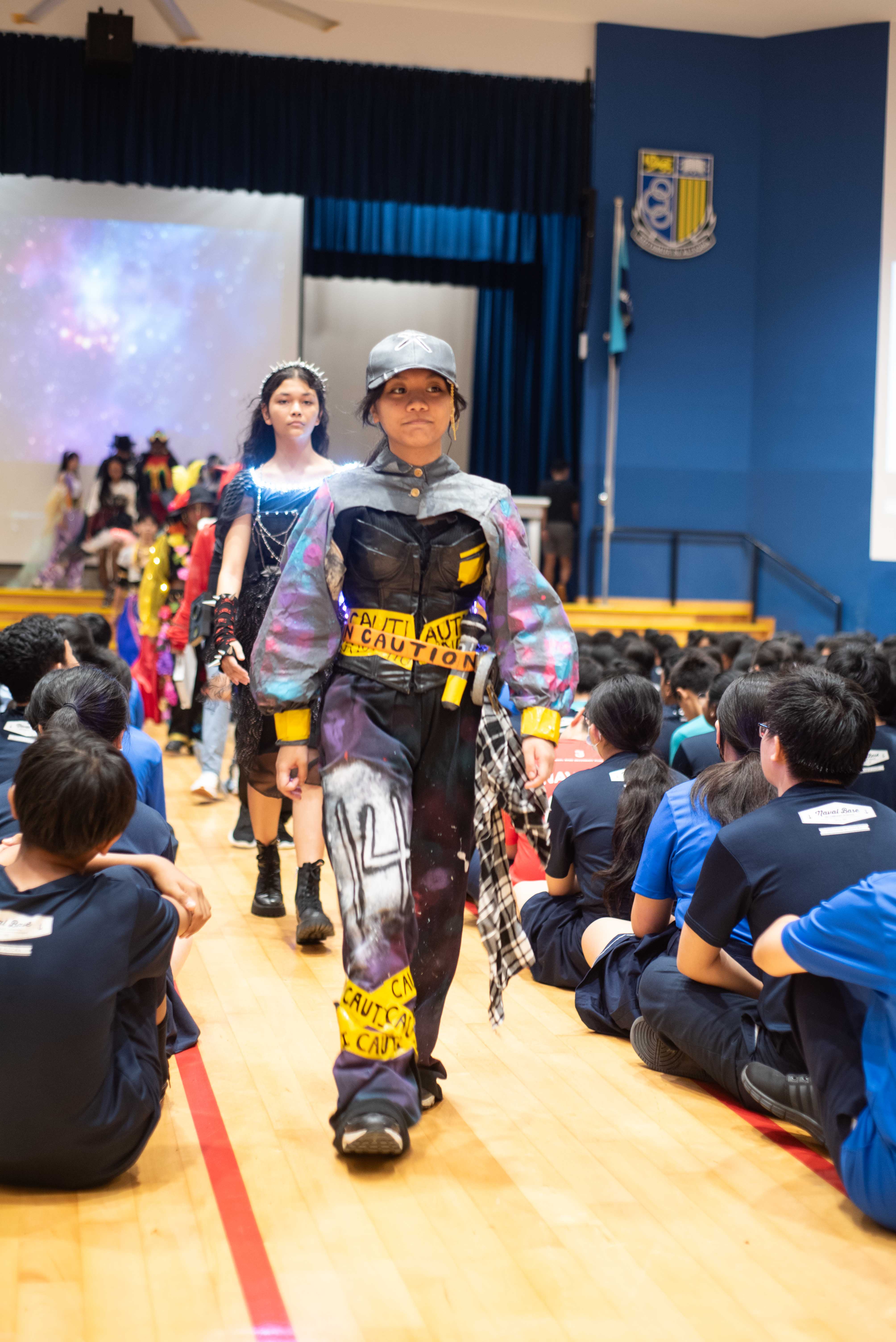 Students walk a runway in creative outfits with caution tape details as seated students watch in a school hall.