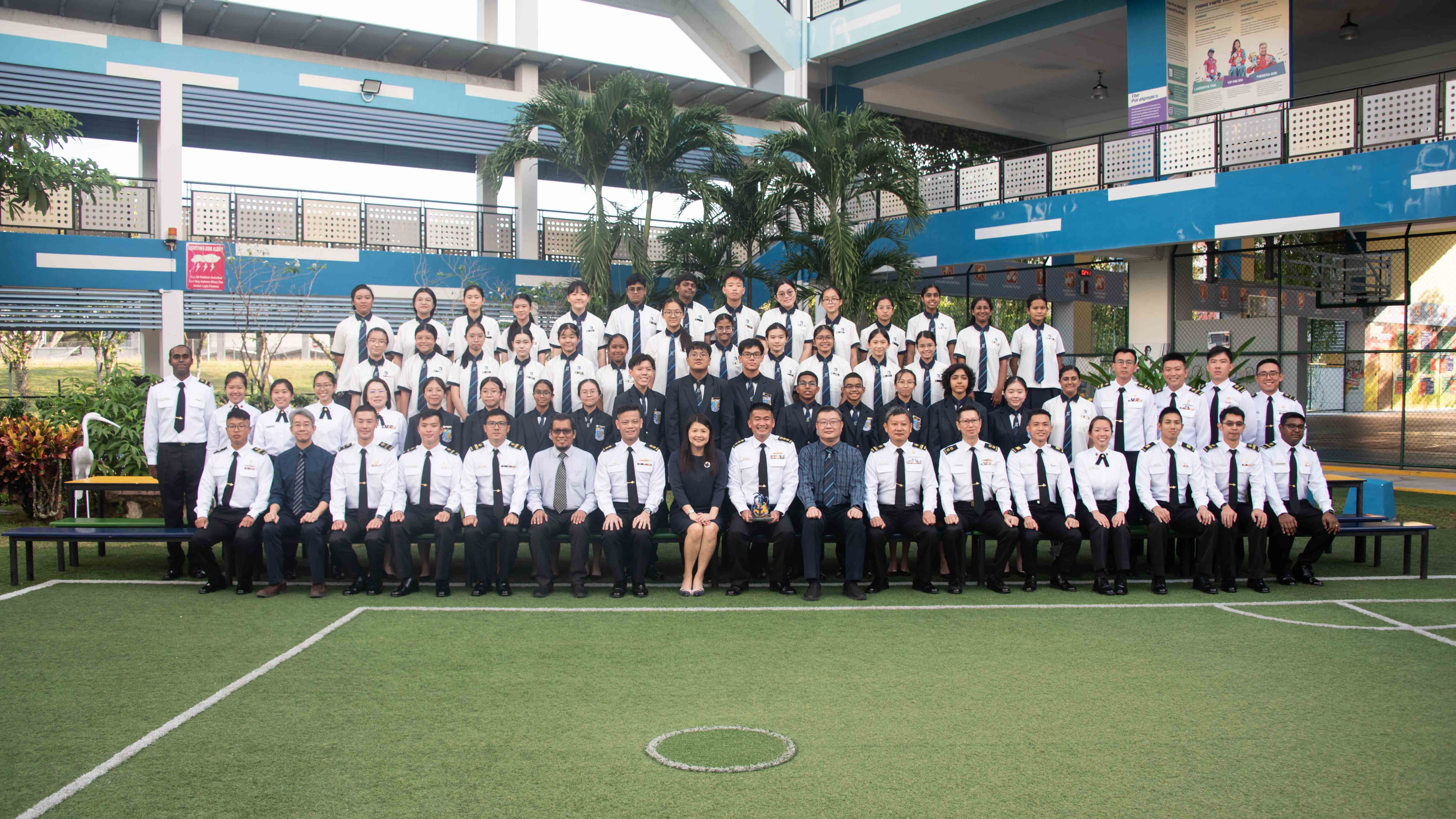 Large group of uniformed students and staff posing for a formal photo on a school field with blue buildings behind.