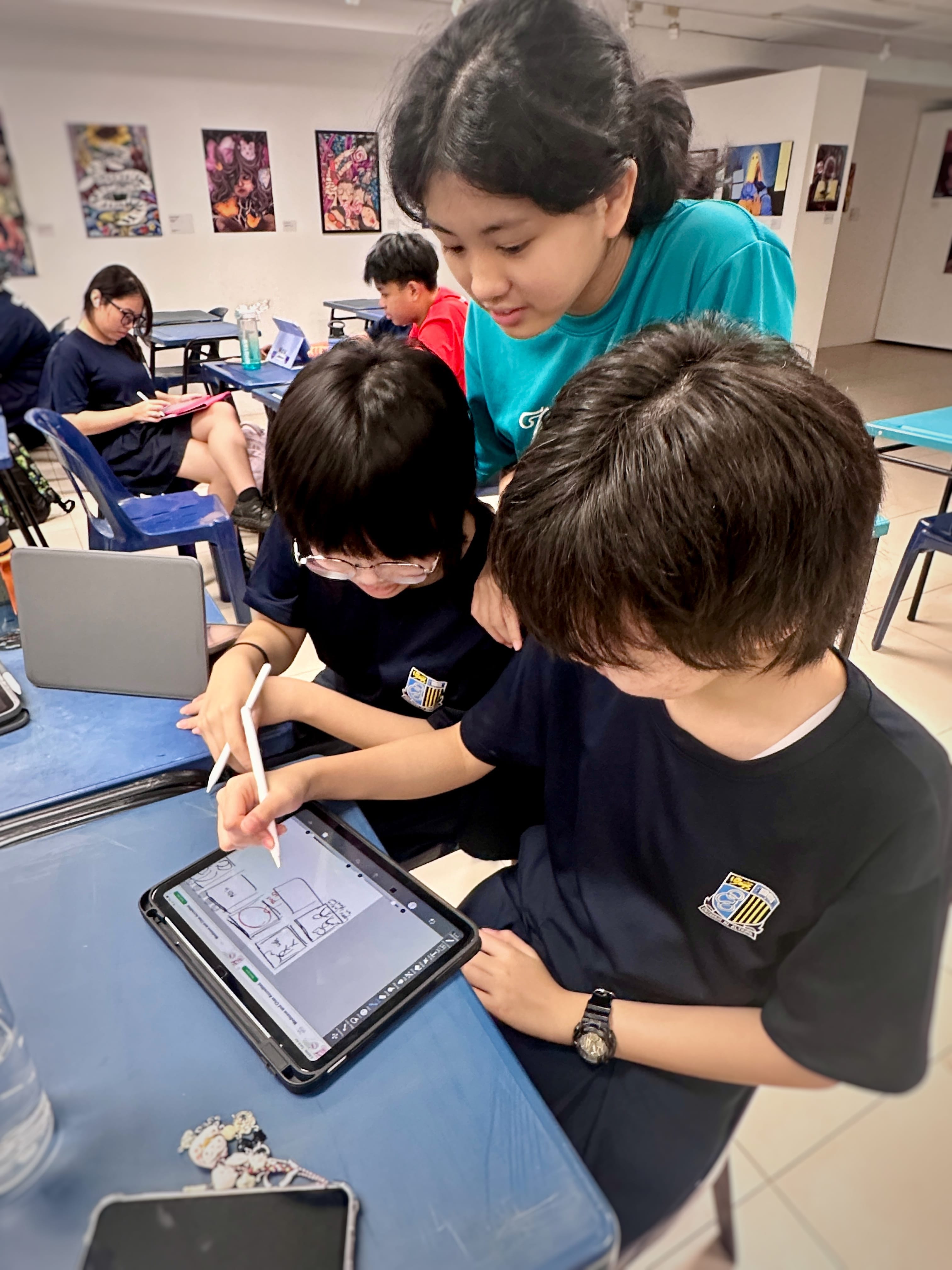 Three students in a classroom huddle over a tablet, one drawing with a stylus while colorful artwork hangs on the walls behind them.
