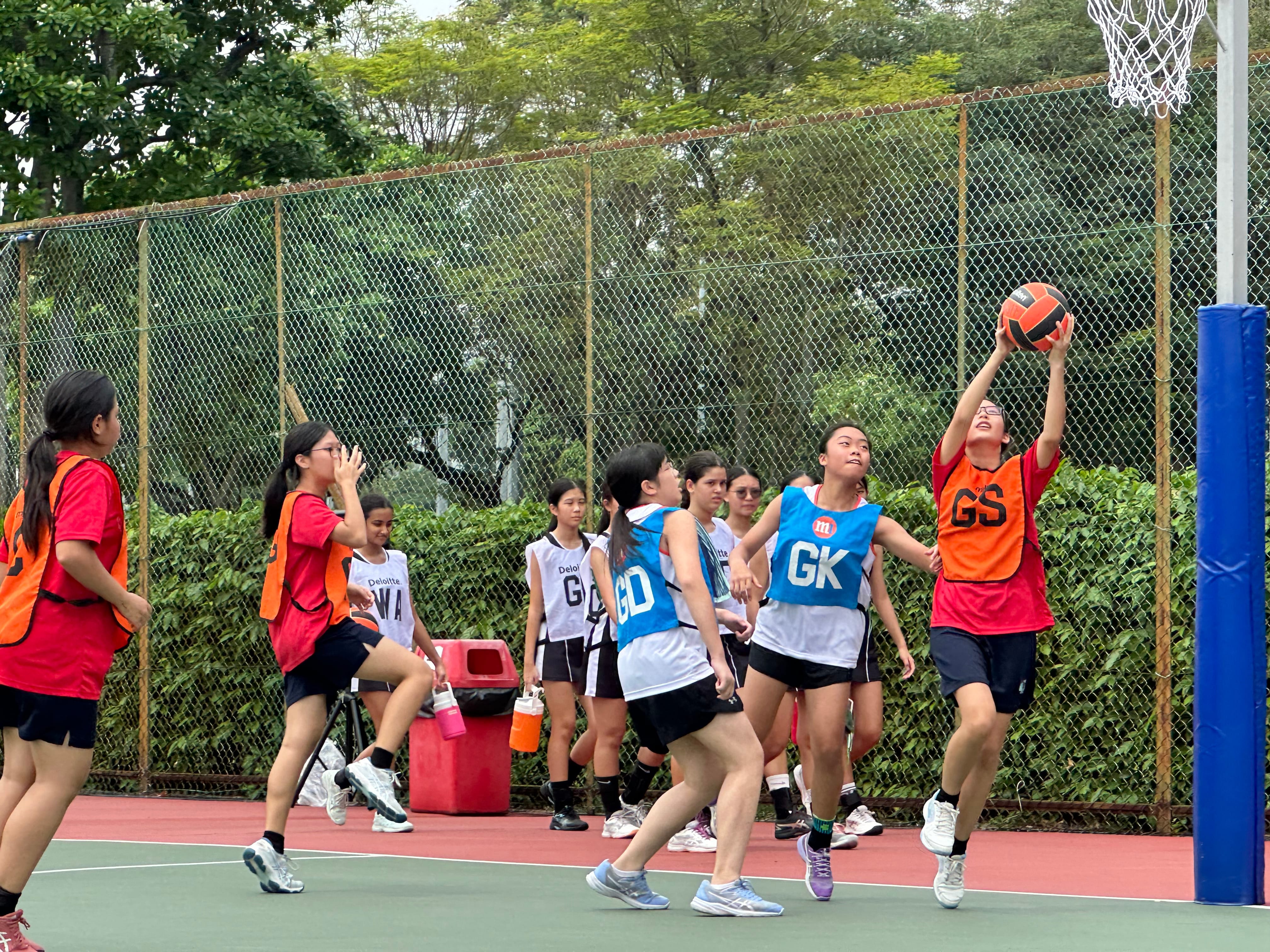 Girls playing netball; shooter in orange jumps to catch ball near hoop as defenders in blue reach up.