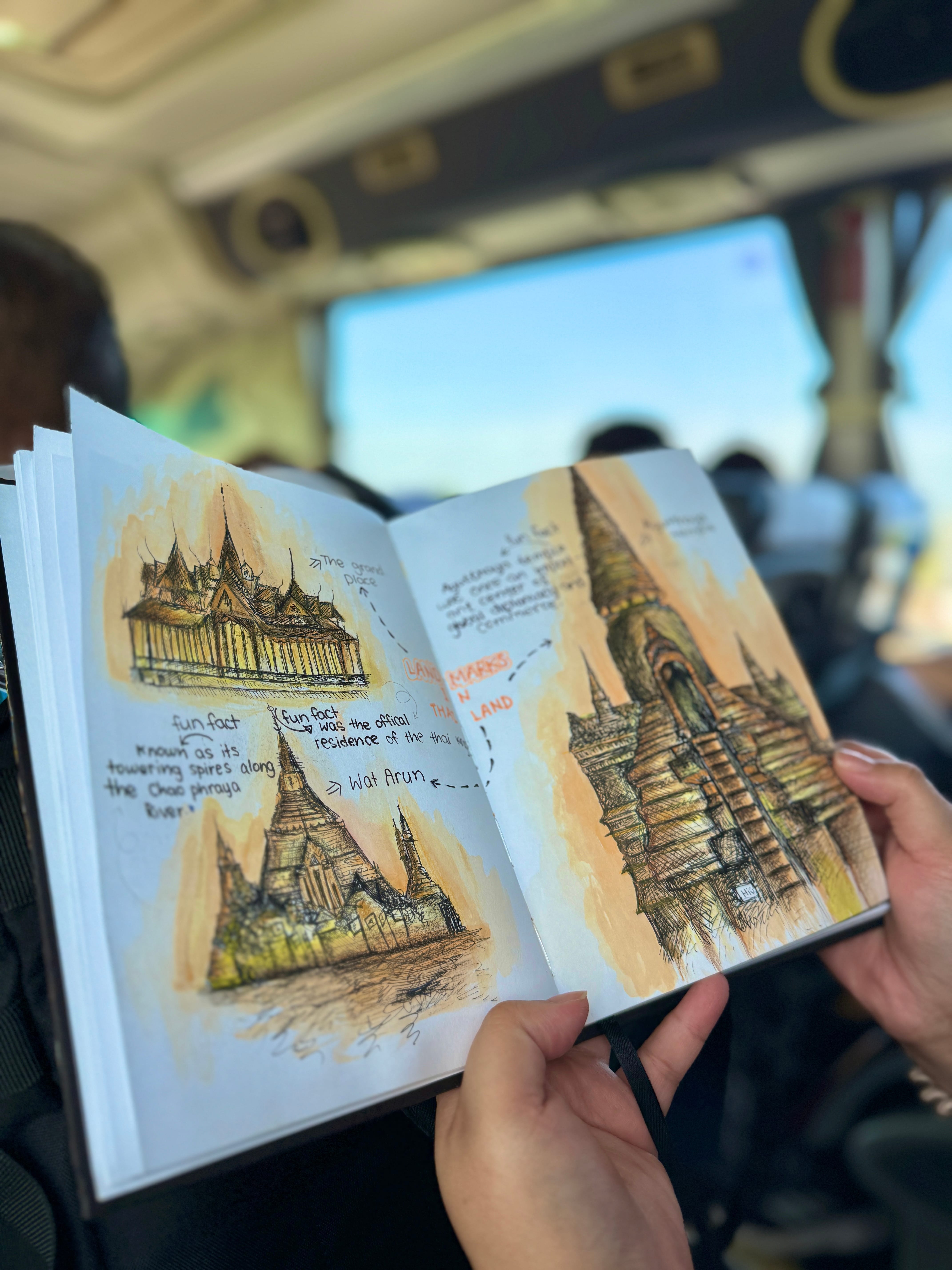 Hands hold an open sketchbook with drawings of temple spires, on a bus with passengers blurred behind.