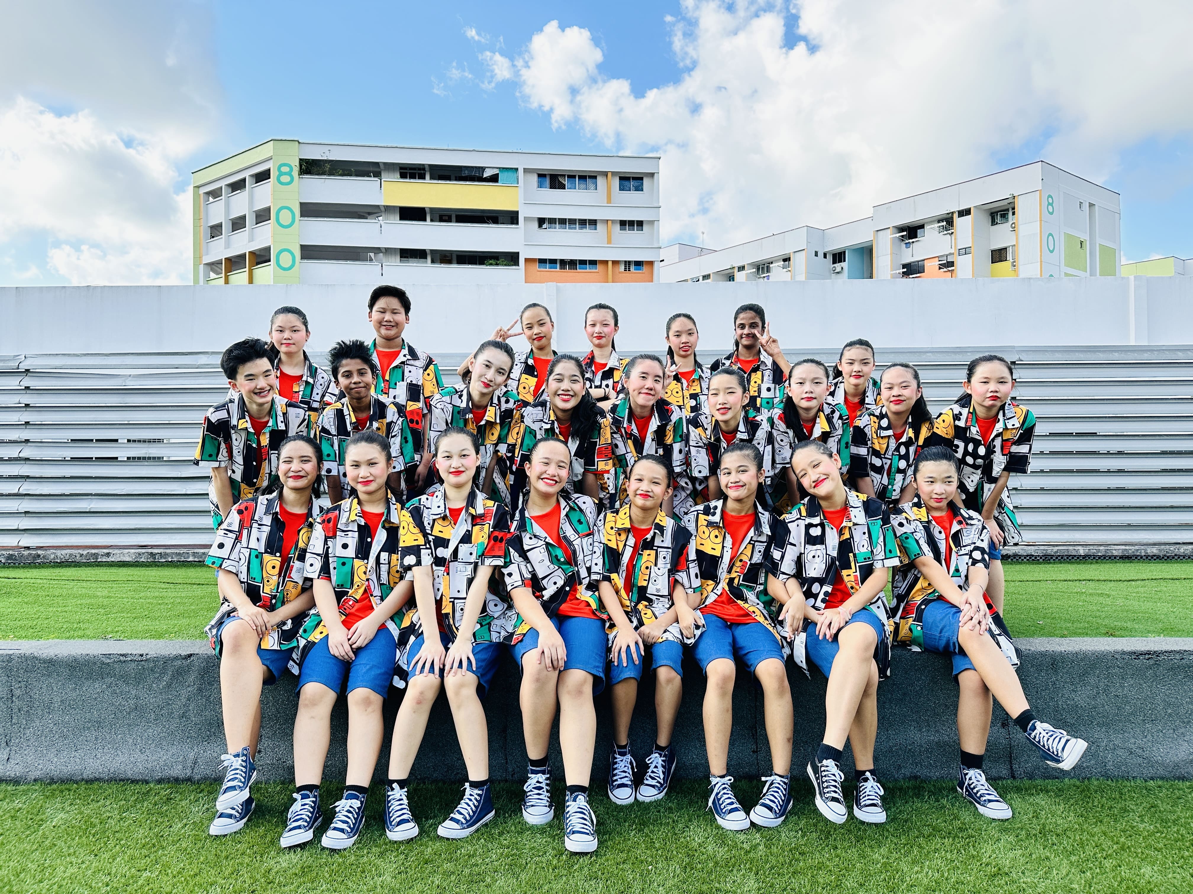 A group of children in matching colorful patterned shirts, red tops, and blue shorts pose on stadium bleachers outdoors.