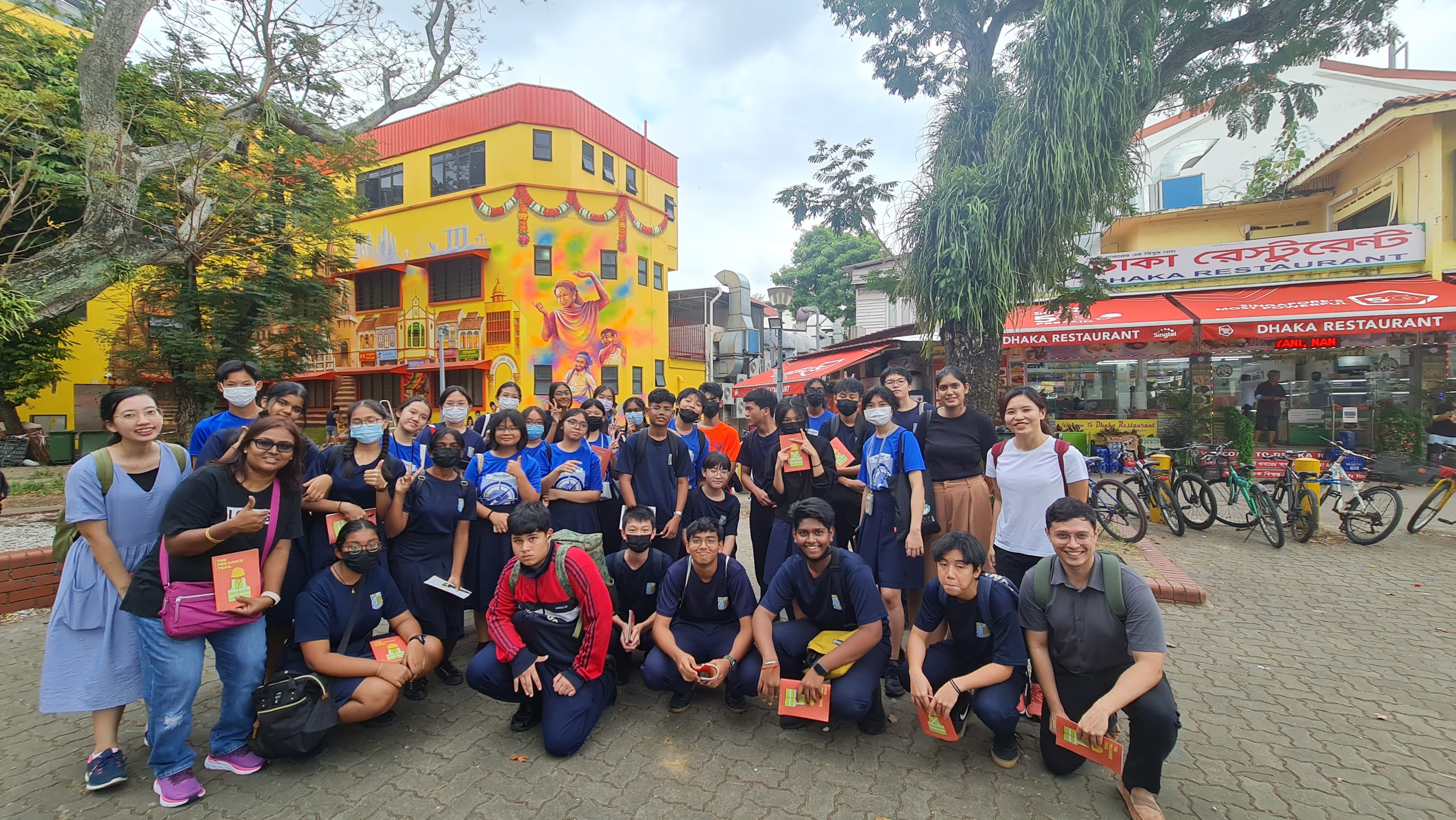 A large group of students in blue uniforms pose outdoors near a colorful mural building and Dhaka Restaurant sign.