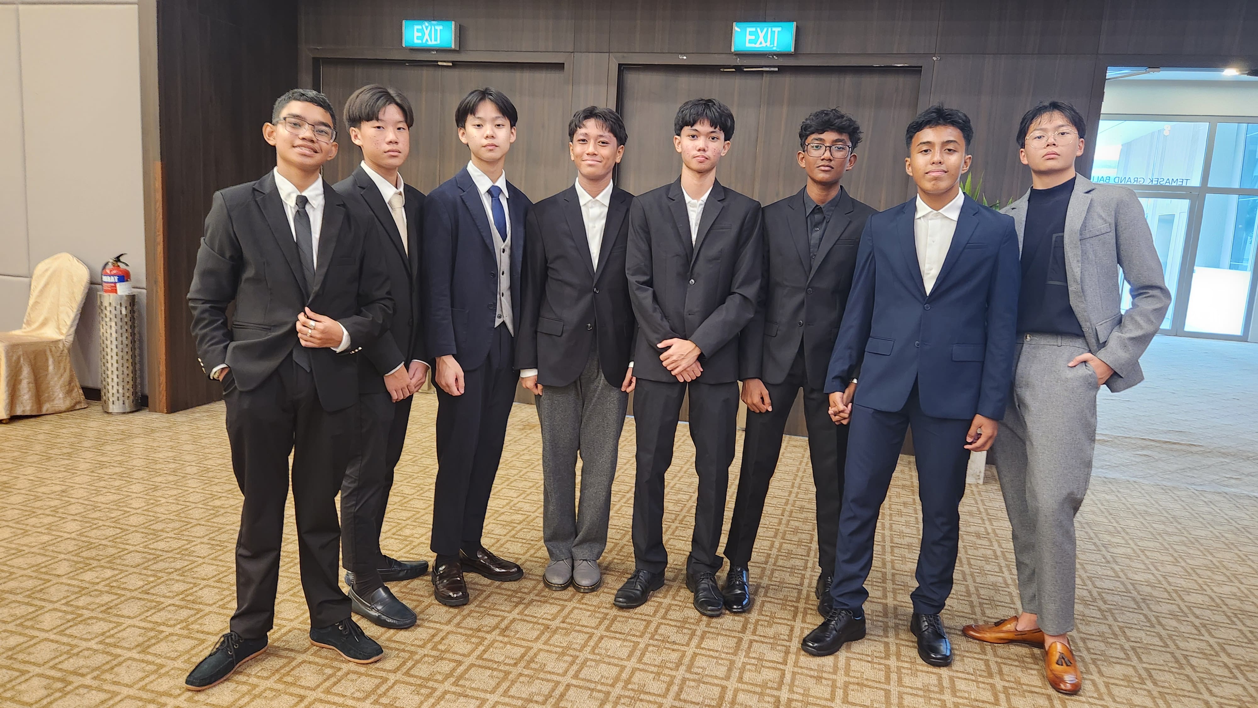Eight young men in formal suits and blazers stand together in a row inside a hall with exit signs.