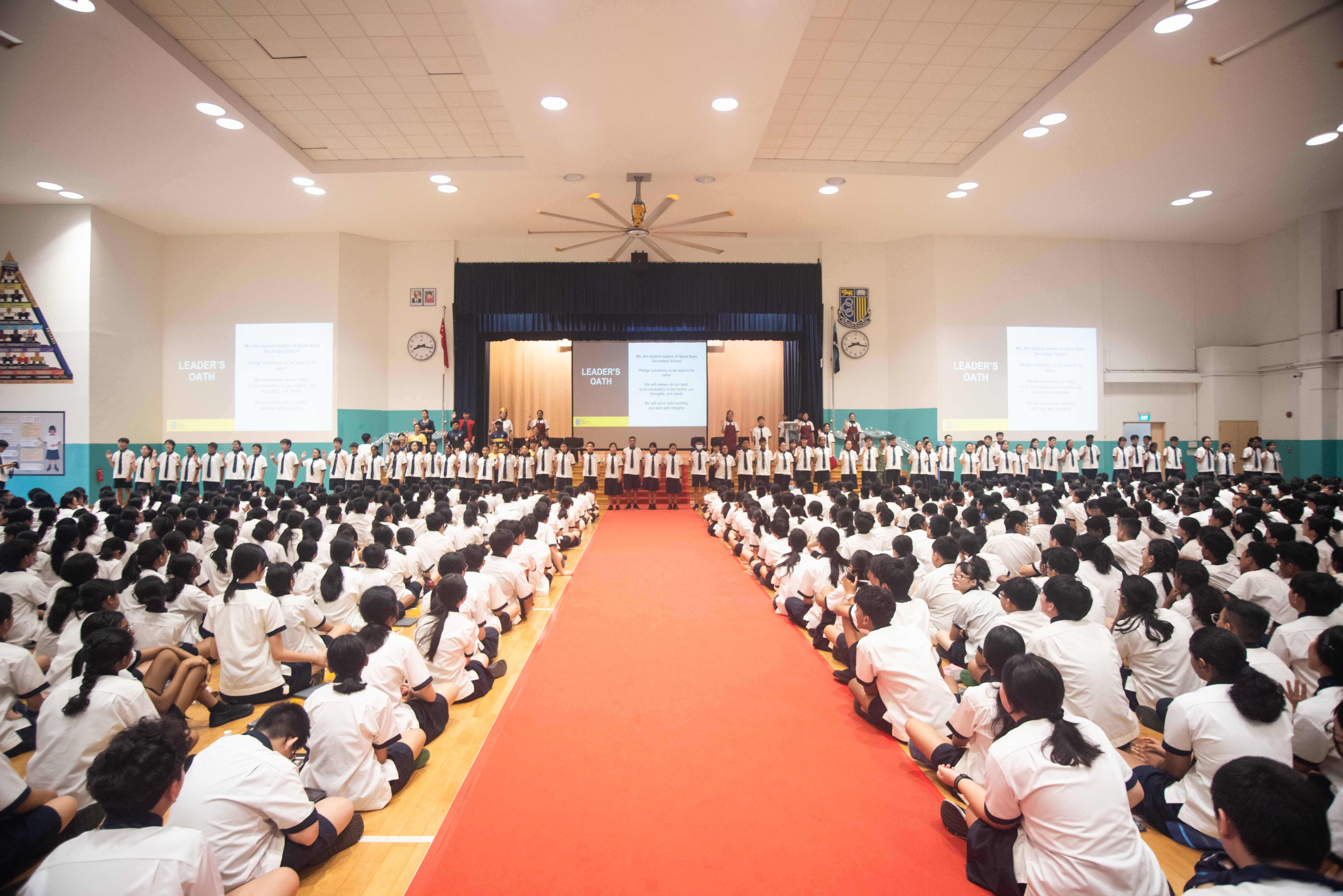 Hundreds of students in white uniforms sit on a gym floor during a Leader's Oath ceremony with a red carpet aisle.