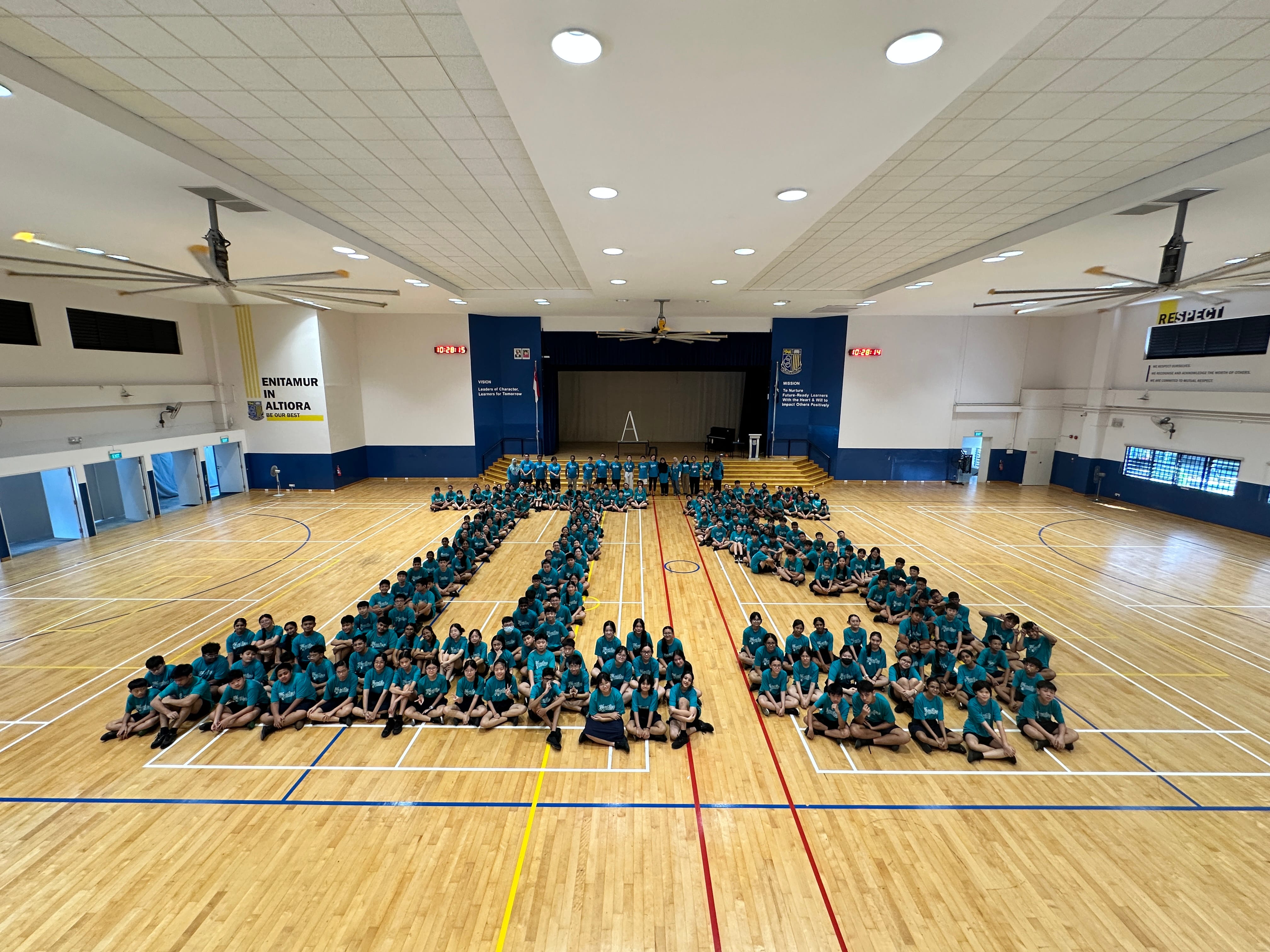 Hundreds of students in teal shirts arranged in a number shape on a school gymnasium wooden floor.