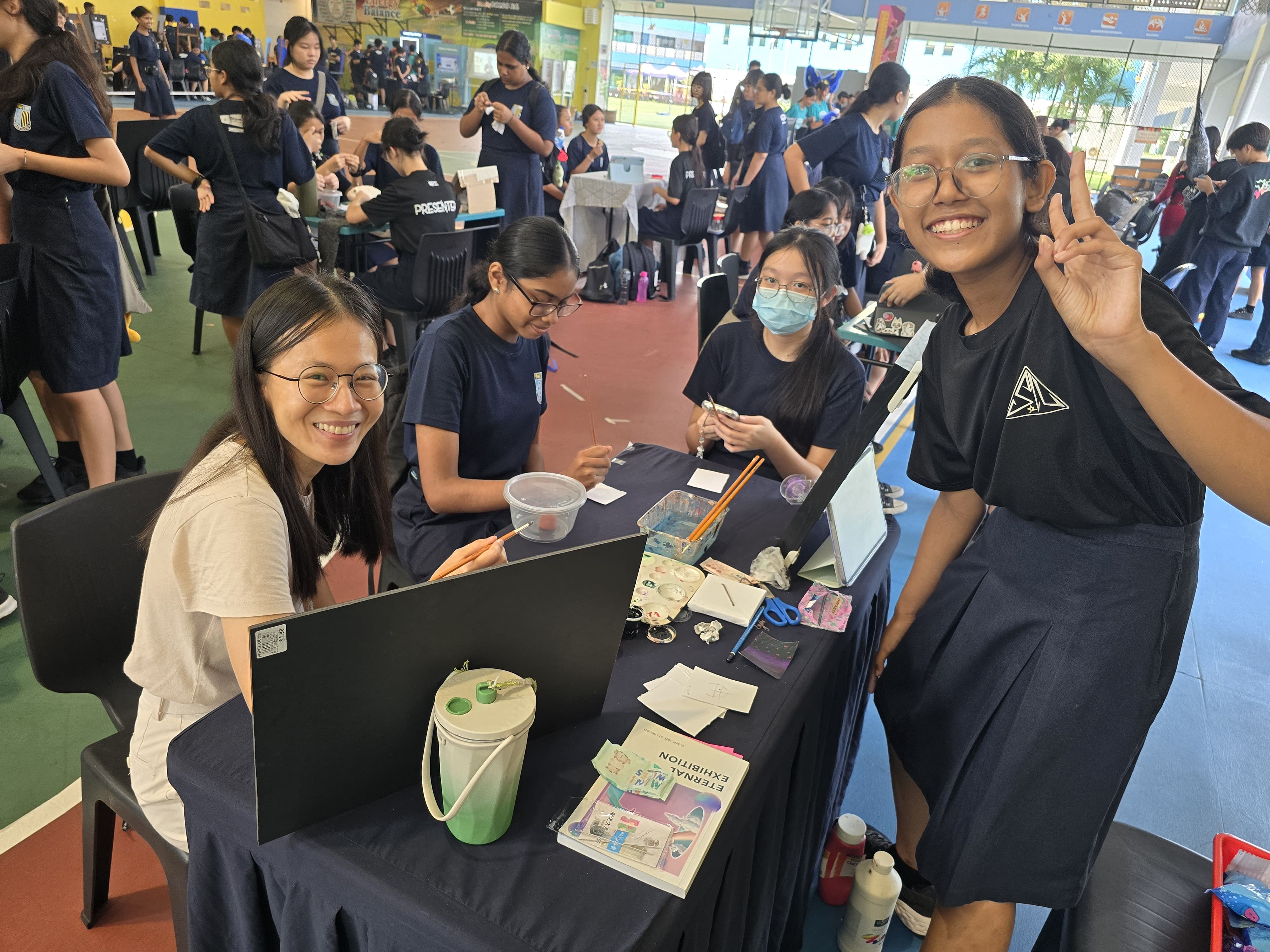 Students in dark uniforms smile at a school event booth with craft supplies and a booklet on the table.
