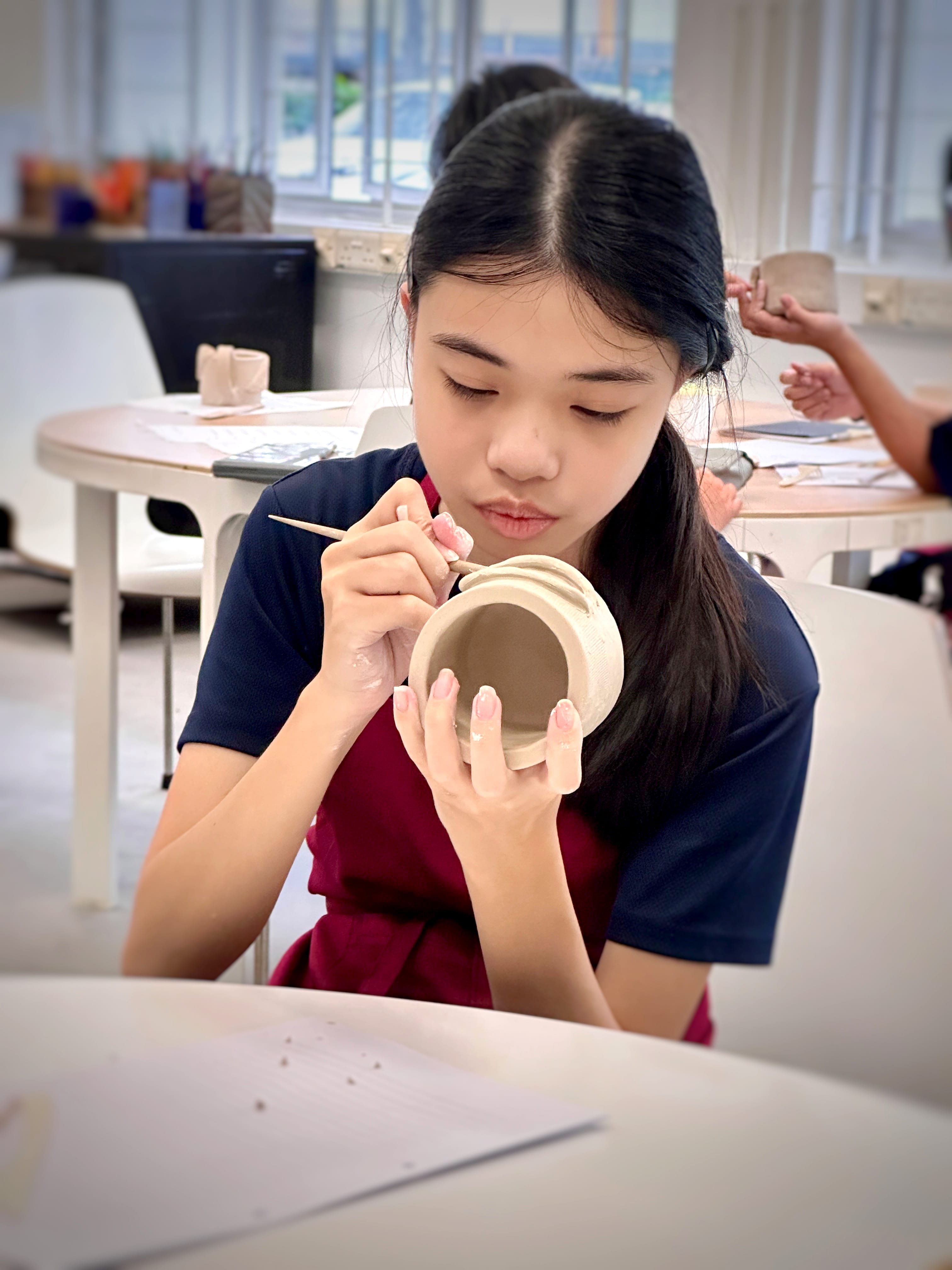 A young girl focuses closely while using a tool to detail a small clay pot in a classroom setting.