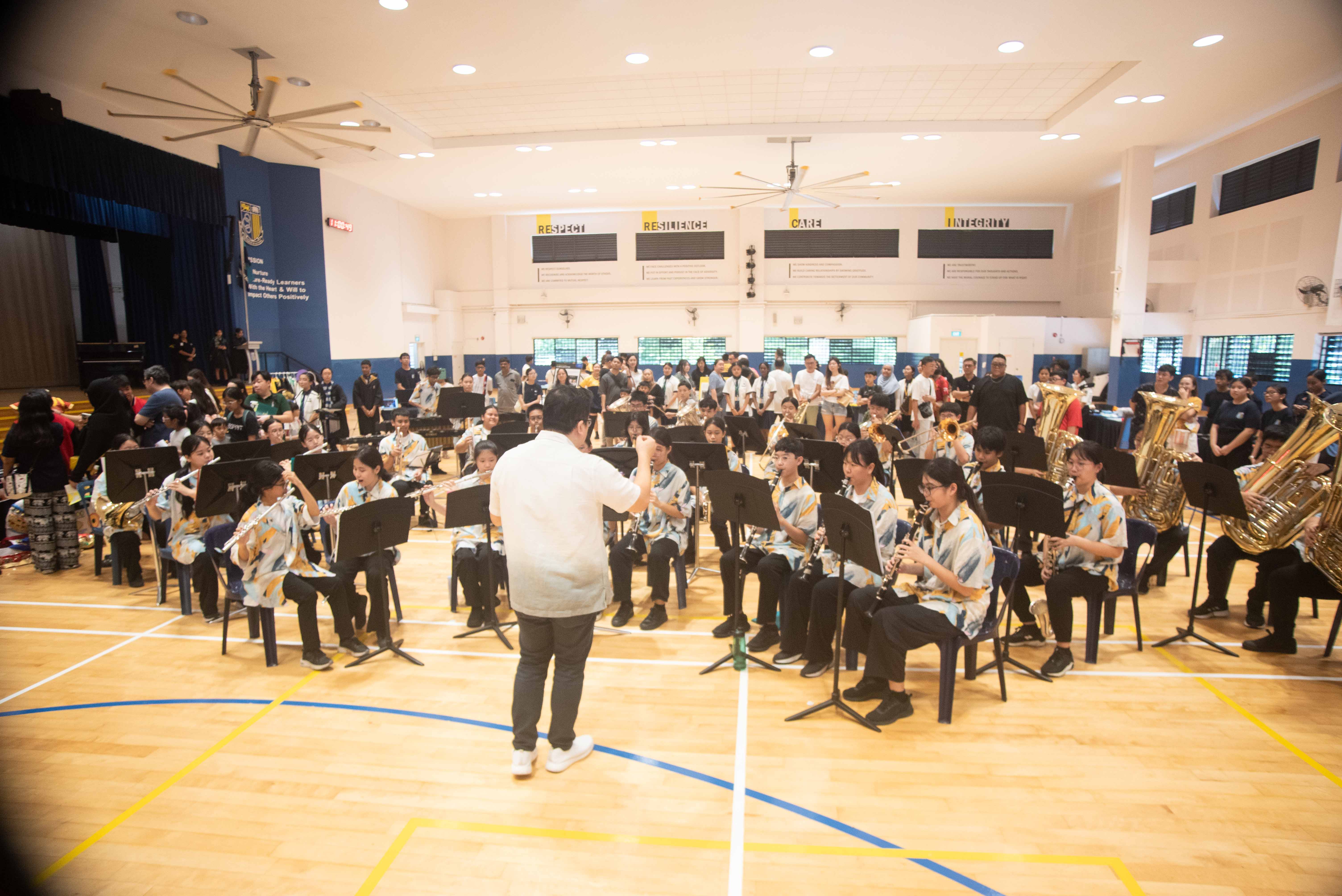 A conductor leads a school band of students playing wind instruments in a gymnasium with an audience watching.