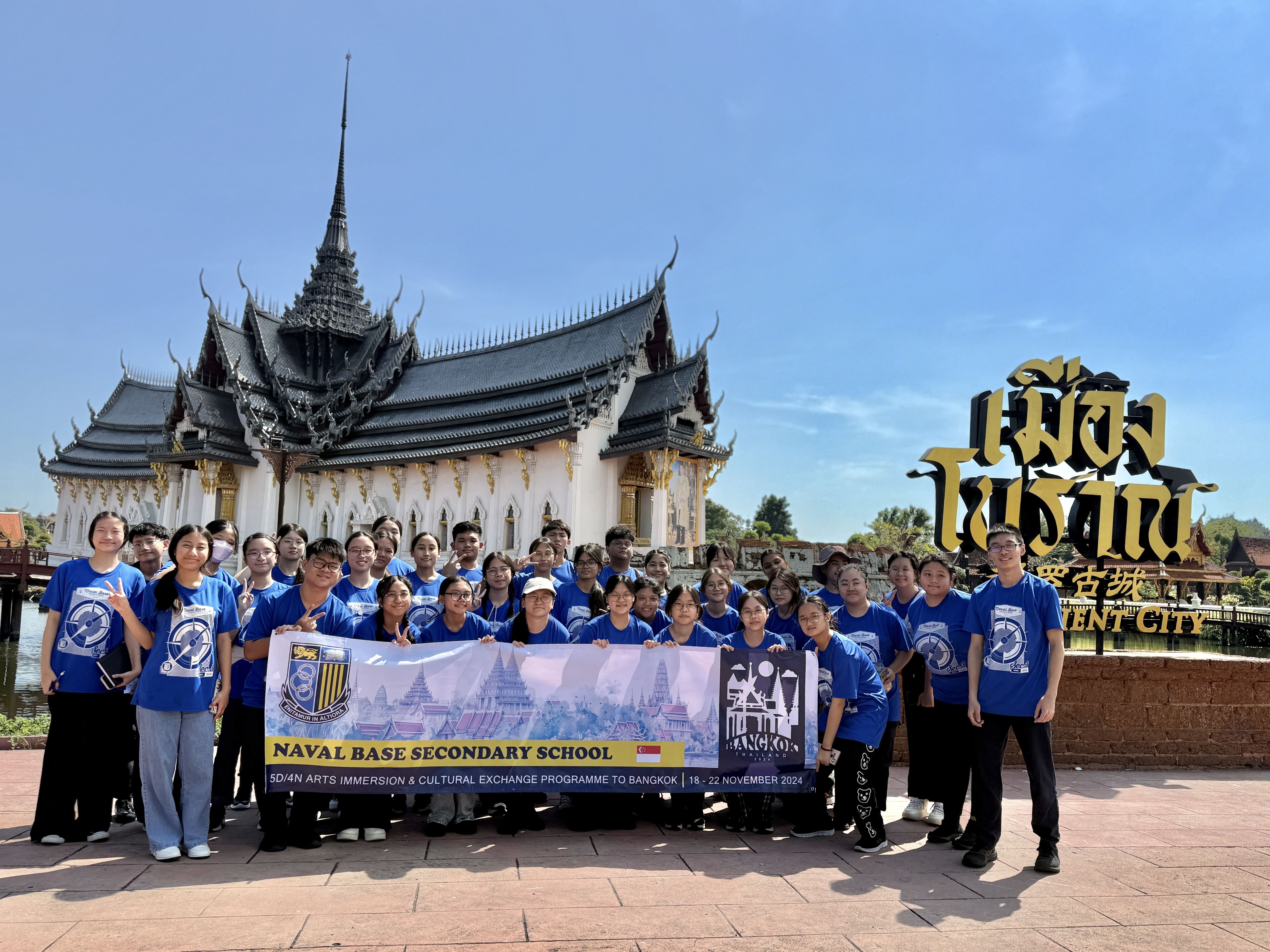 Students in blue shirts hold a Naval Base Secondary School Bangkok trip banner in front of a Thai temple.