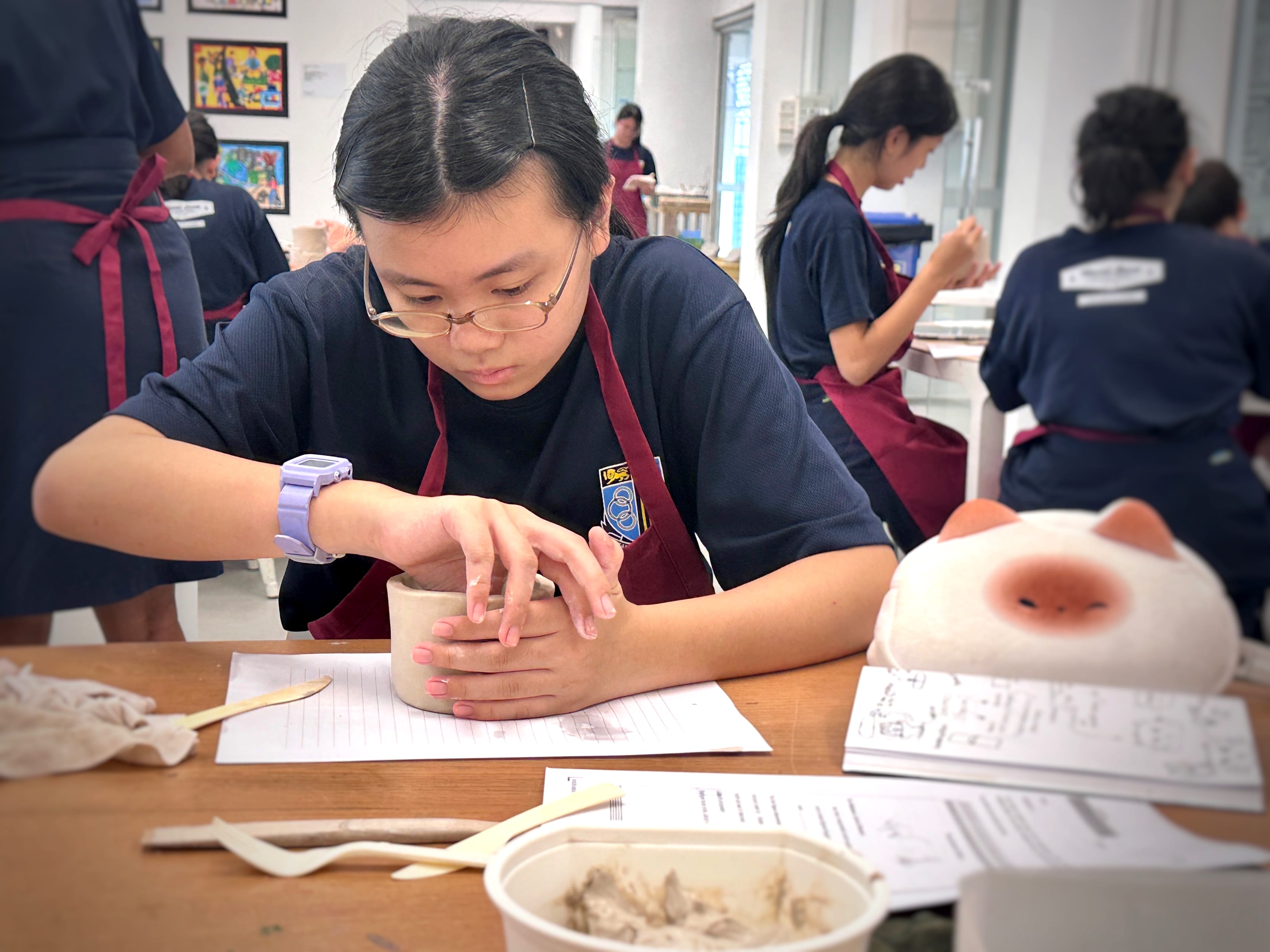 A young student in a navy uniform and burgundy apron shapes a clay pot by hand at a table in an art classroom.