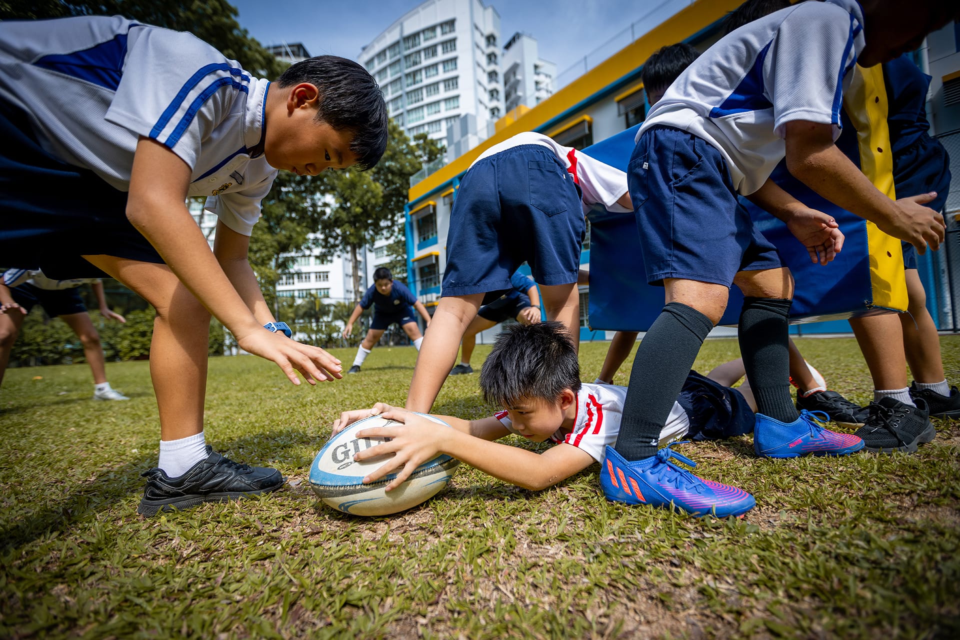Boys in PE uniforms compete for a rugby ball on a grass field, with one boy lying on the ground gripping it.