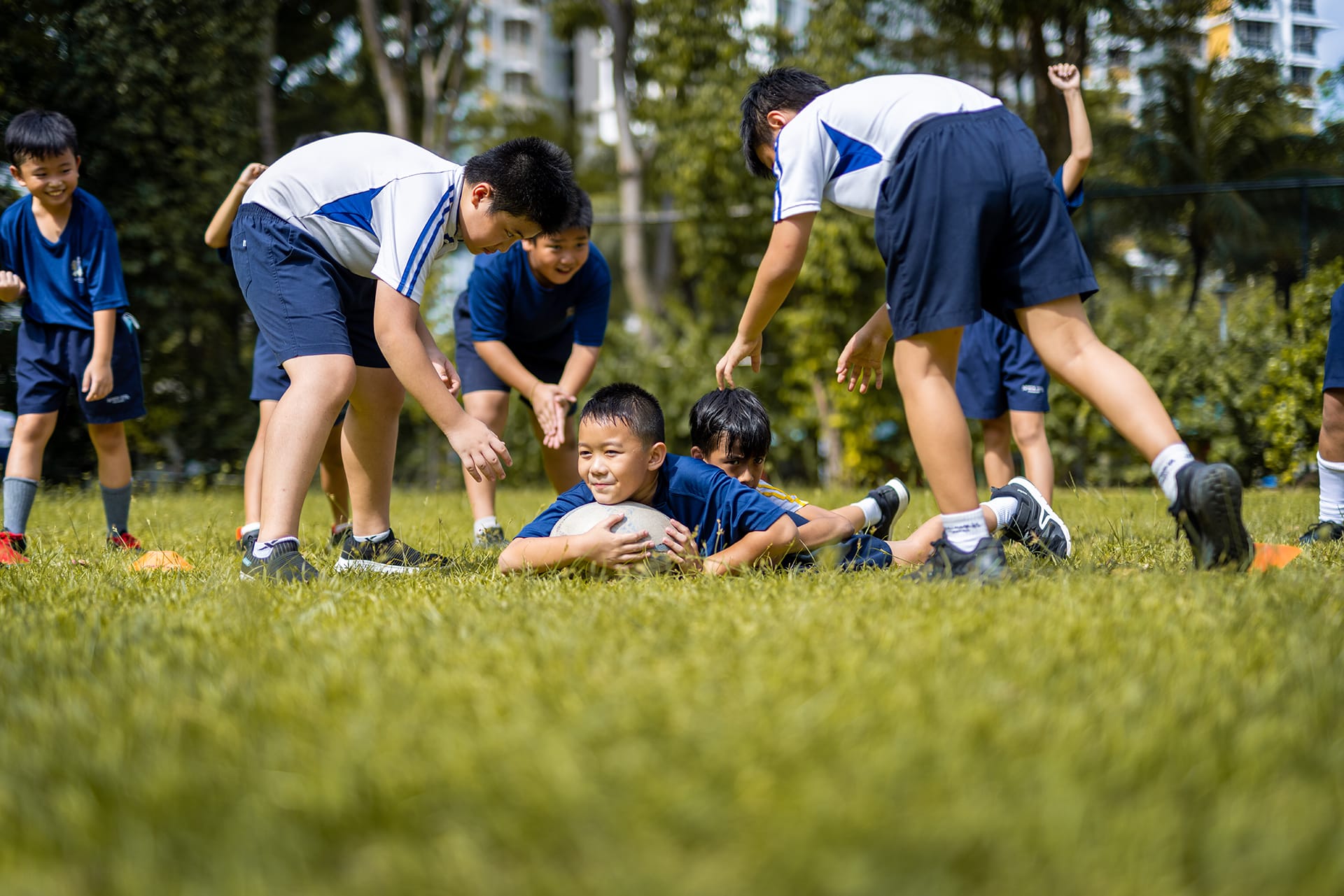 A boy lies on grass clutching a rugby ball while several other boys in uniforms surround him outdoors.