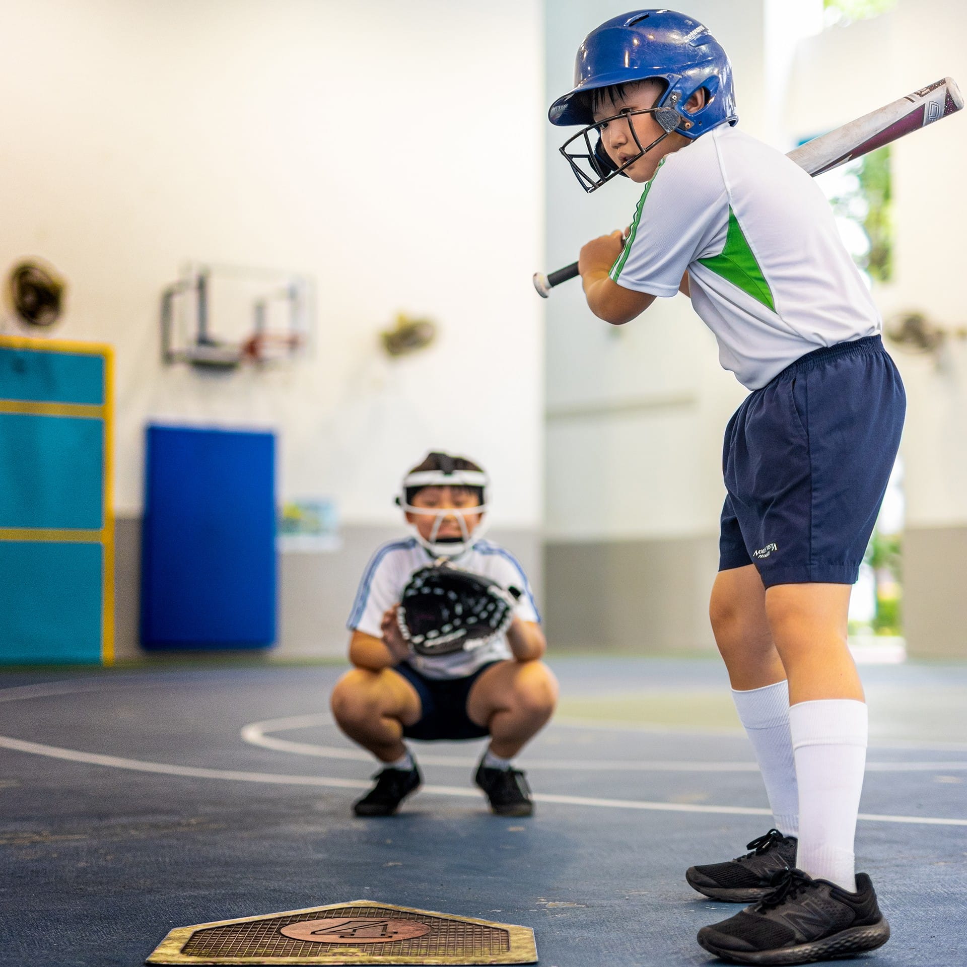 Young boy in a blue helmet and white uniform holds a bat at home plate indoors, with a catcher crouching behind him.