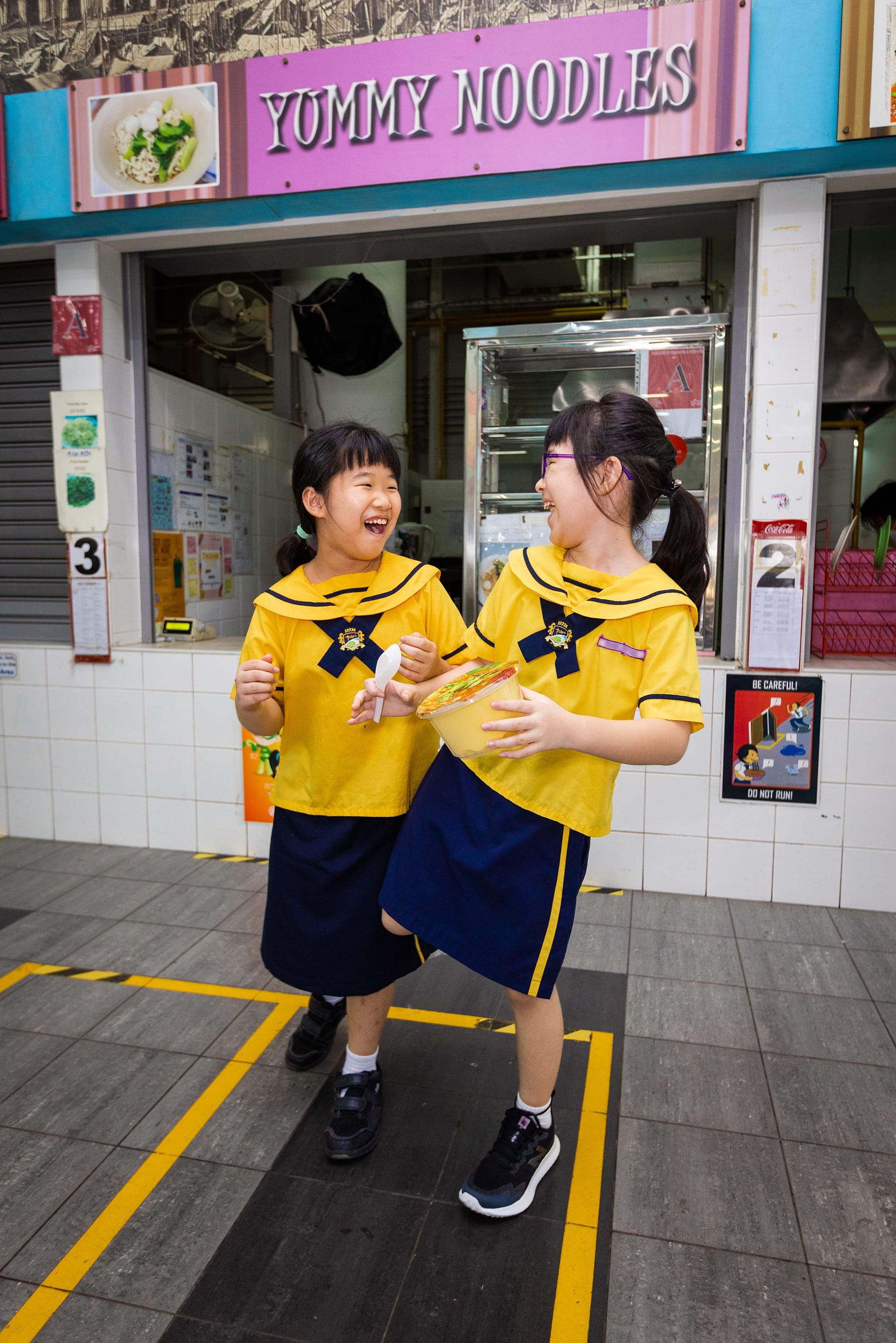 Two schoolgirls in yellow uniforms laugh outside a stall under a Yummy Noodles sign, holding a bowl.