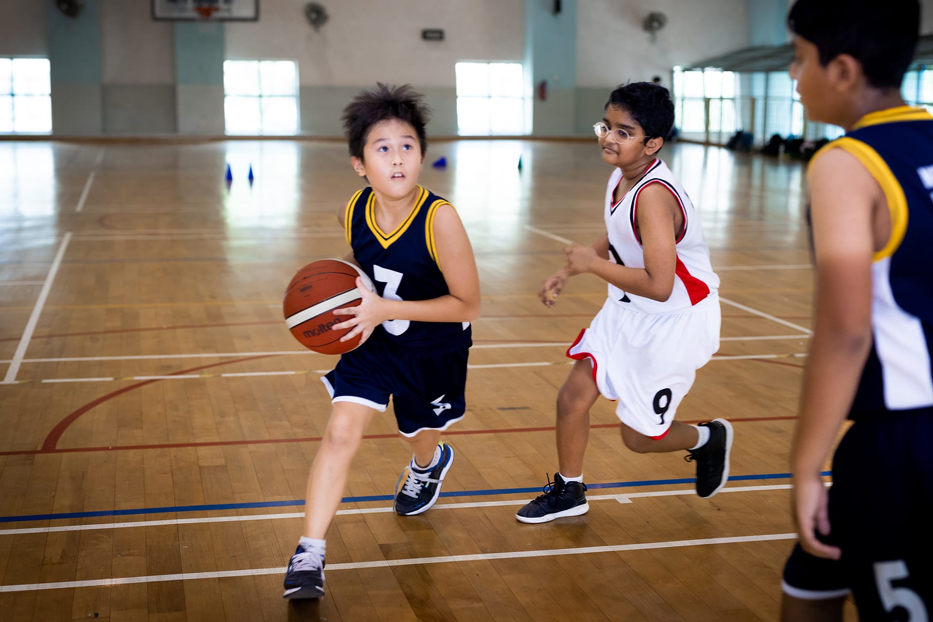 3 young boys playing basketball indoors, 1 in navy holding the ball while the other in white chases after him to block his advance.