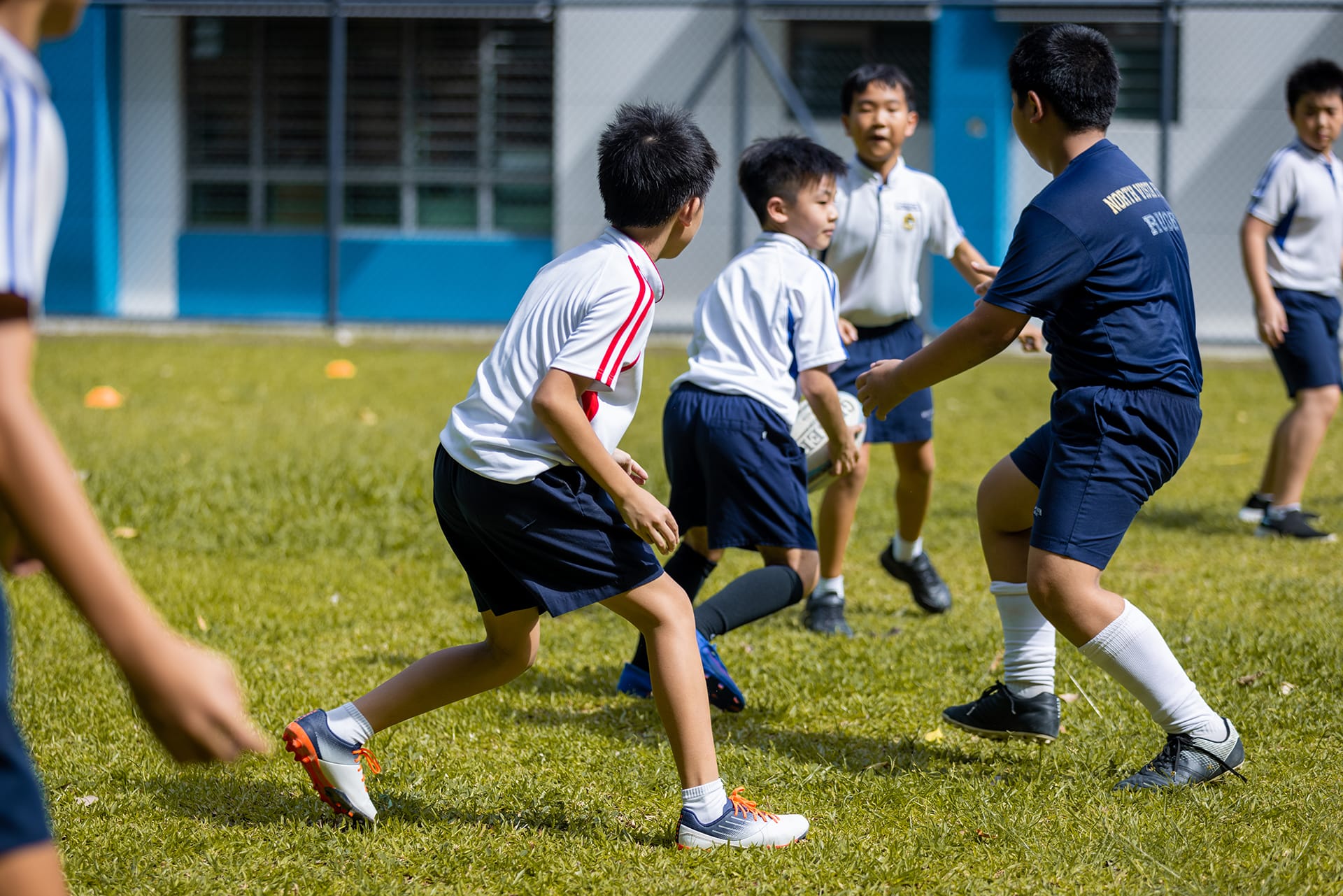 Several young boys in white and navy uniforms play rugby on a grass field outside a school building.