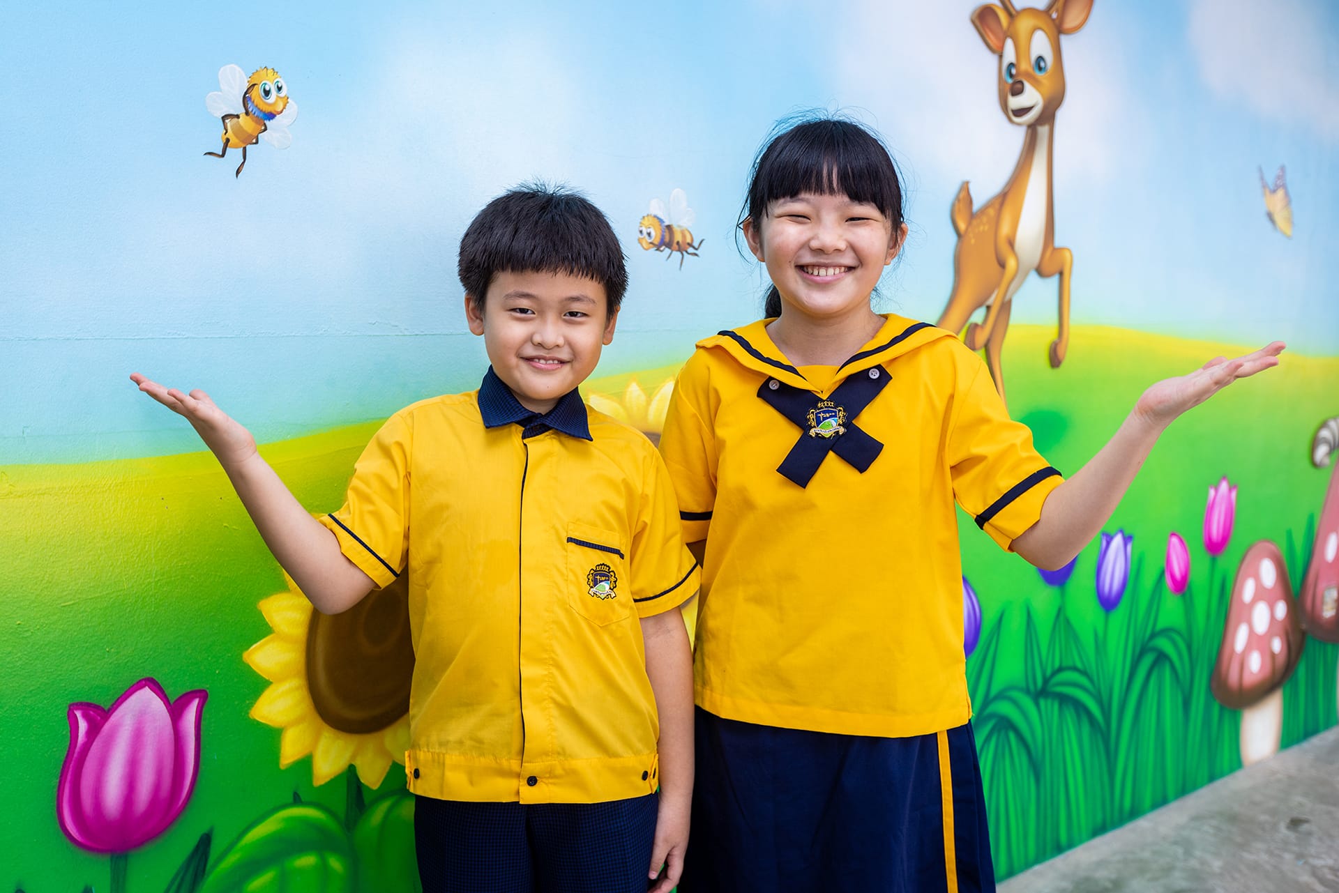 Two smiling children in yellow school uniforms stand in front of a colorful mural with a deer, bees, and flowers.