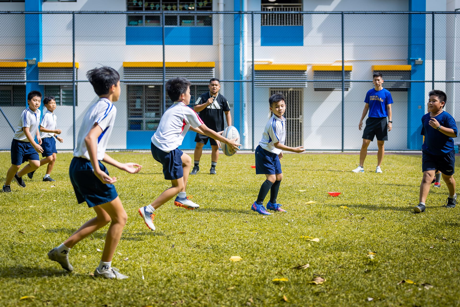 Young boys in white uniforms play rugby on a grass field near a blue and white school building, supervised by two adults.