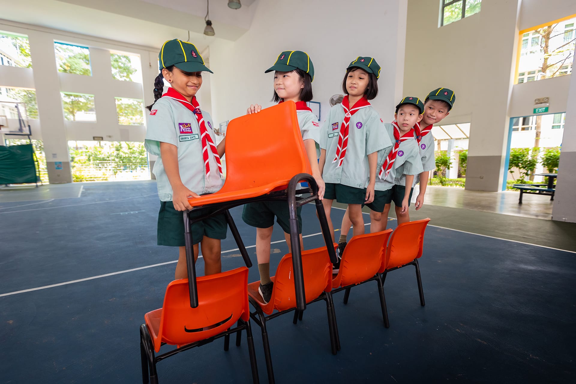 Five children in scout uniforms with green caps and red scarves carry orange plastic chairs across an indoor court.