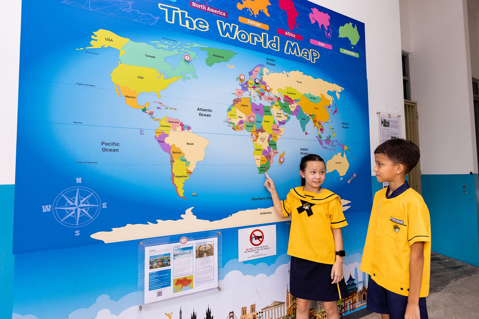 Two children in yellow uniforms stand before a large colorful world map mural, with a girl pointing at it.