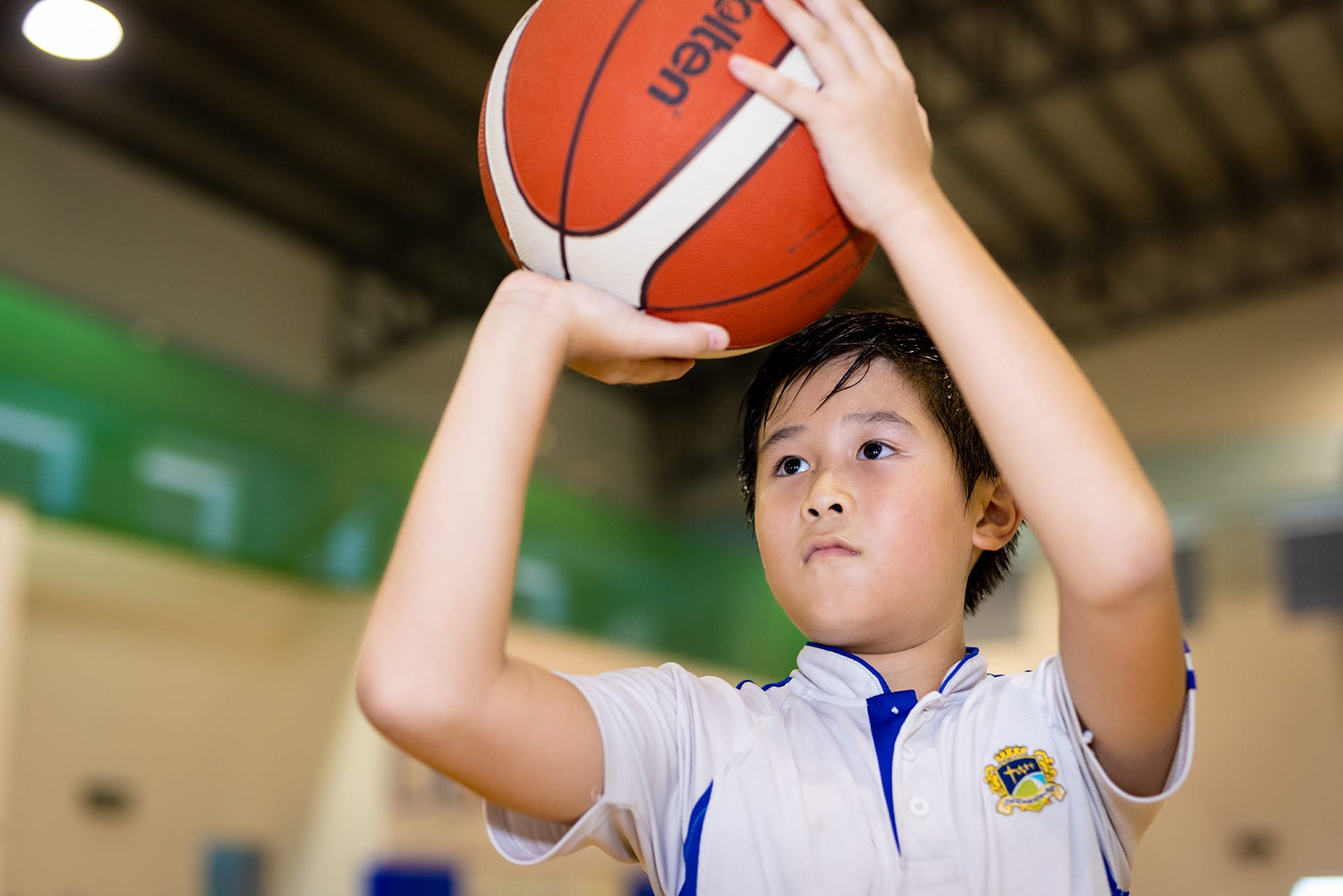 A young boy in a white and blue uniform holds a Molten basketball above his head, preparing to shoot indoors.
