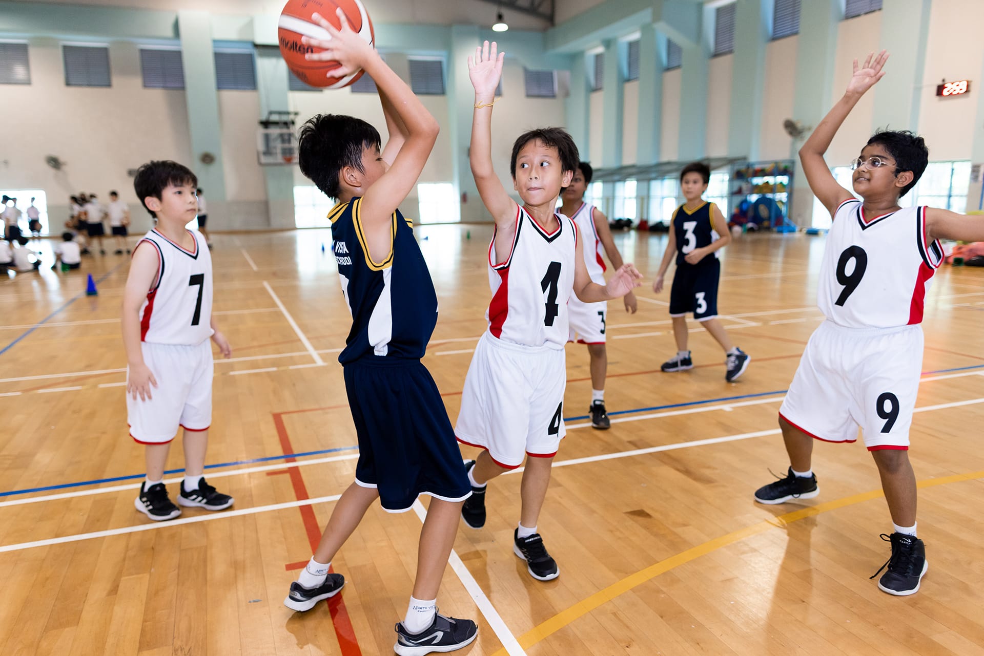 Young children in numbered jerseys play basketball indoors, one boy attempts to pass to his teammates while defenders raise their arms to block his pass.