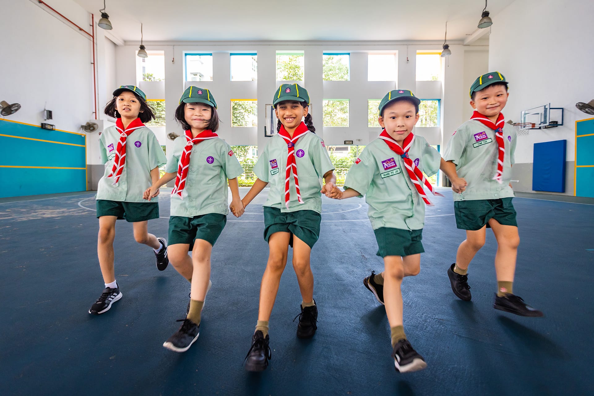 Five children in scout uniforms with green caps and red scarves march holding hands inside the school indoor sports all.