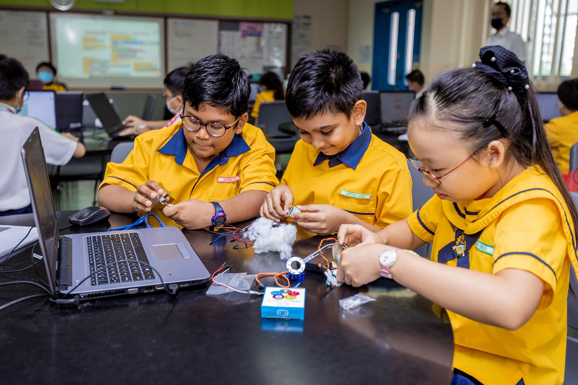 Three children in yellow uniforms build an electronics project with wires and components at a desk beside a laptop.