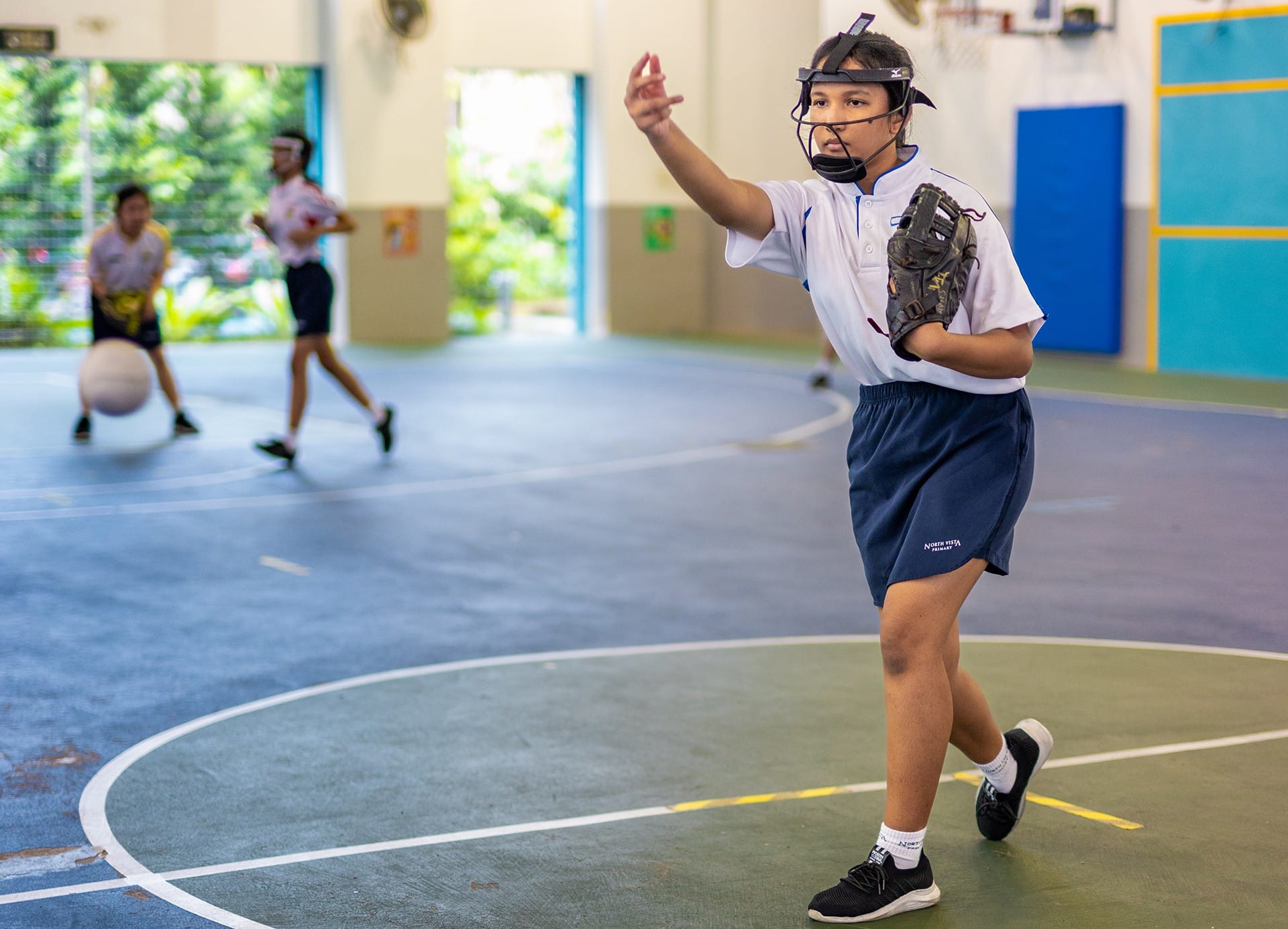 A child wearing a face guard and baseball glove prepares to throw in an indoor sports hall.