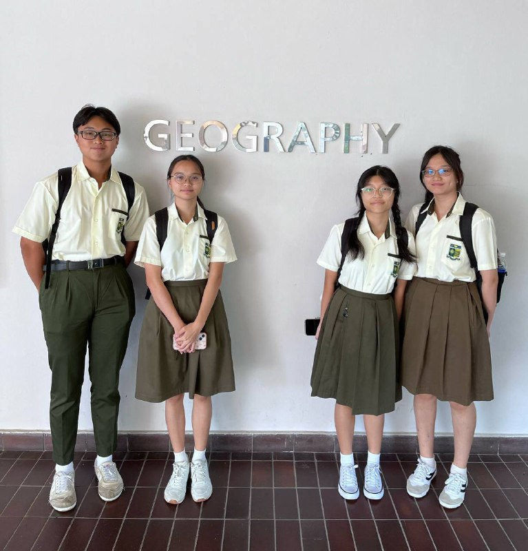 Four students in matching green and white uniforms stand in front of a wall with the word GEOGRAPHY.