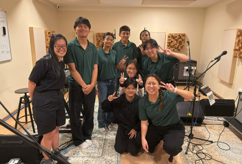 A group of nine smiling students and youth pose together on a decorative rug inside a music recording studio. Most of them are wearing matching dark green polo shirts, and several are making peace signs at the camera. The studio features sound-dampening wooden wall panels, microphones on stands, a keyboard, and audio amplifiers in the background.