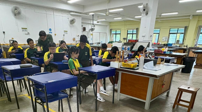 A classroom setting where students wearing green and yellow school uniforms are seated at blue desks, working on personal laptop tablets. A male teacher in a green shirt leans over to assist a student in the back row. In the foreground, another student stands by a desk to collaborate with a classmate. The background shows an expansive design and technology workshop with large windows and heavy workbenches.
