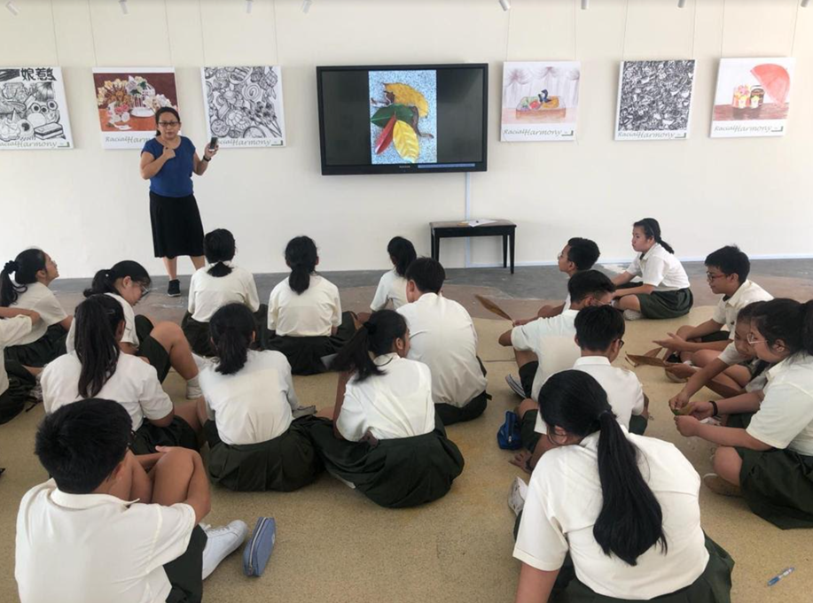 A female teacher stands at the front of a room, speaking to a group of students sitting cross-legged on the floor in a semi-circle. A large TV screen behind the teacher displays an image of colorful leaves. The wall is decorated with square art prints under the heading "Racial Harmony," and the students are wearing school uniforms consisting of white shirts and dark skirts or trousers.