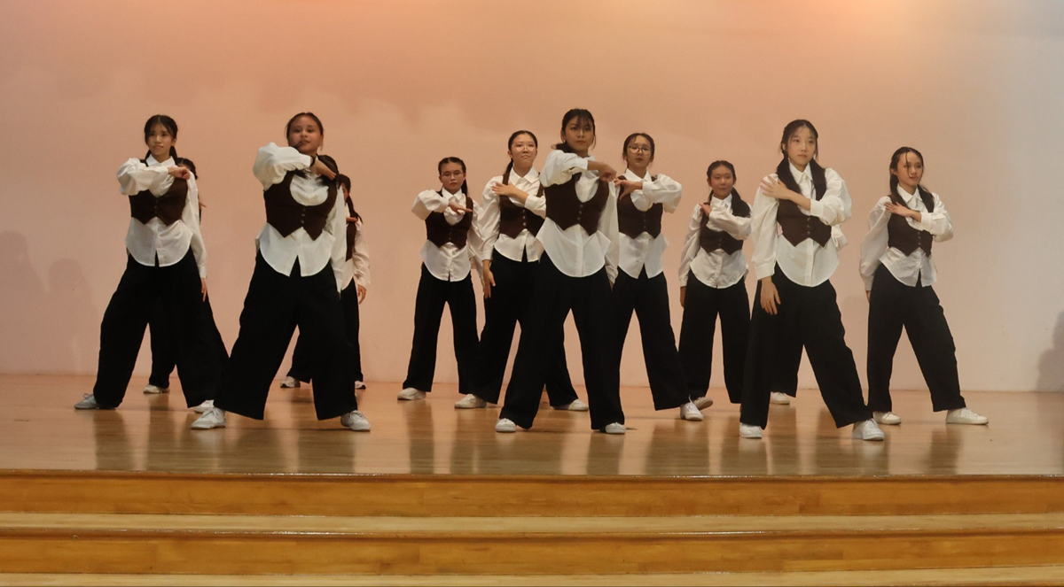 A group of young female dancers performing a synchronized routine on a wooden stage. They are wearing matching outfits consisting of white long-sleeved collared shirts, dark brown vests, loose-fitting black wide-leg trousers, and white sneakers, set against a plain peach-colored background.