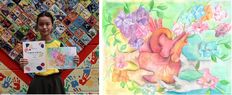 A girl holds a certificate and artwork beside a colorful mural; the artwork shows a heart surrounded by flowers.