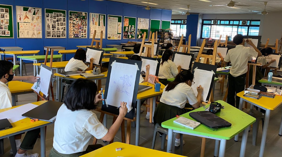 Students in school uniforms are seated at bright yellow and green desks in a spacious art studio, working diligently on sketches mounted on wooden table easels. The classroom features blue accent walls lined with student artwork displays, and large windows are visible in the background.