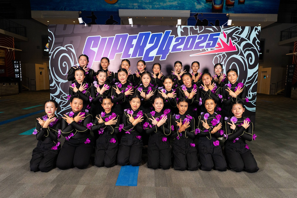 A group photo of a large female dance crew posing in front of a graphic backdrop that reads "SUPER 24 2025". The dancers are arranged in rows wearing matching black long-sleeved outfits decorated with purple orchid flowers, all crossing their arms over their chests in a uniform pose.