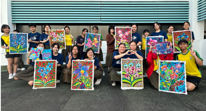 A group photo of students standing outdoors against a white building wall, proudly holding up their completed, brightly colored batik paintings. Most of the artworks feature intricate floral and orchid designs with vivid blue, green, red, and yellow patterns. The students are smiling, with some kneeling in the front row and others standing behind them.