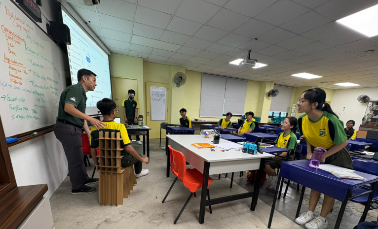 An interactive classroom session where a teacher in a green polo shirt stands near a whiteboard filled with notes, gesturing toward a student sitting on a unique cardboard prototype chair. A digital projector screen displays a presentation in the background. Other students, dressed in yellow and green uniforms, watch attentively from their blue desks arranged around a central white table.