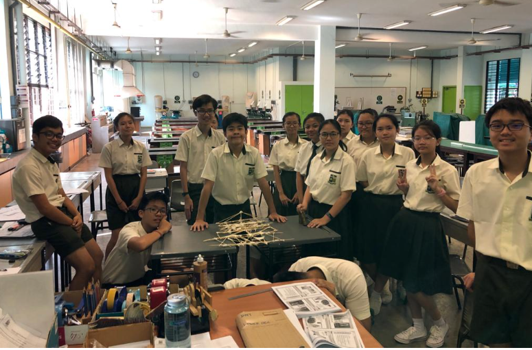 A group of about twelve secondary school students in white short-sleeved shirts and dark skirts or shorts gather together in a brightly lit school workshop. They are smiling and posing around a table that holds a complex structural prototype made of thin wooden sticks and strings. The large room behind them features long workbenches and machinery.