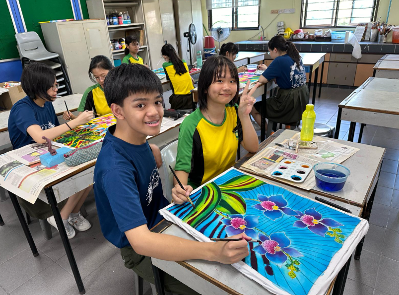 Two smiling students look at the camera while working on batik paintings at their desks in an art classroom. In the foreground, a boy holds a paintbrush over a vibrant blue fabric painting featuring purple orchids and green leaves. Next to him, a girl gives a peace sign. In the background, other students are focused on their own art projects at various desks.