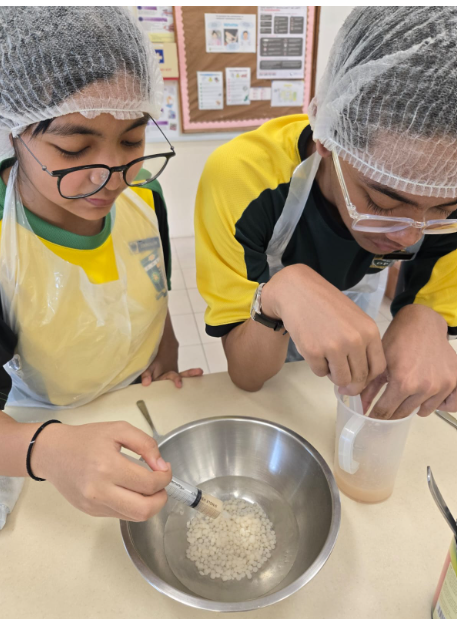 Two students wearing protective hairnets, plastic aprons, and glasses lean intently over a school kitchen counter during a food science experiment. One student uses a syringe to drop a liquid mixture into a stainless steel bowl containing small, white gel-like spheres, while the other student stirs a liquid mixture inside a clear plastic measuring jug next to it.