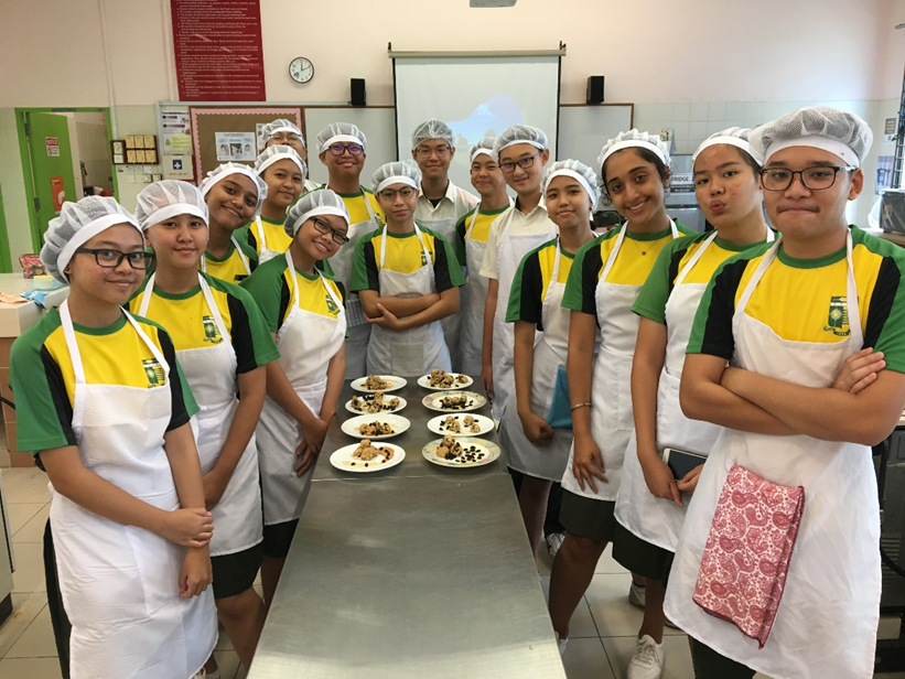 A group photo of fifteen smiling students wearing matching green and yellow school uniforms, white hairnets, and white aprons. They are gathered around a long stainless steel counter in a cookery room, proudly presenting several white plates arranged with small baked treats and decorative toppings.