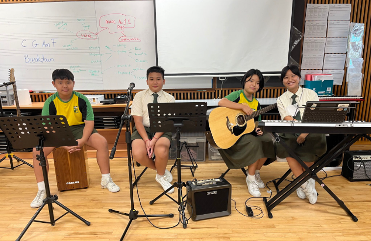 Four secondary school students pose with musical instruments in front of a white classroom board covered in music theory notes. From left to right, a boy sits on a wooden cajon drum, a boy sits behind a microphone stand, a girl holds an acoustic guitar, and another girl sits at an electronic keyboard with a laptop.