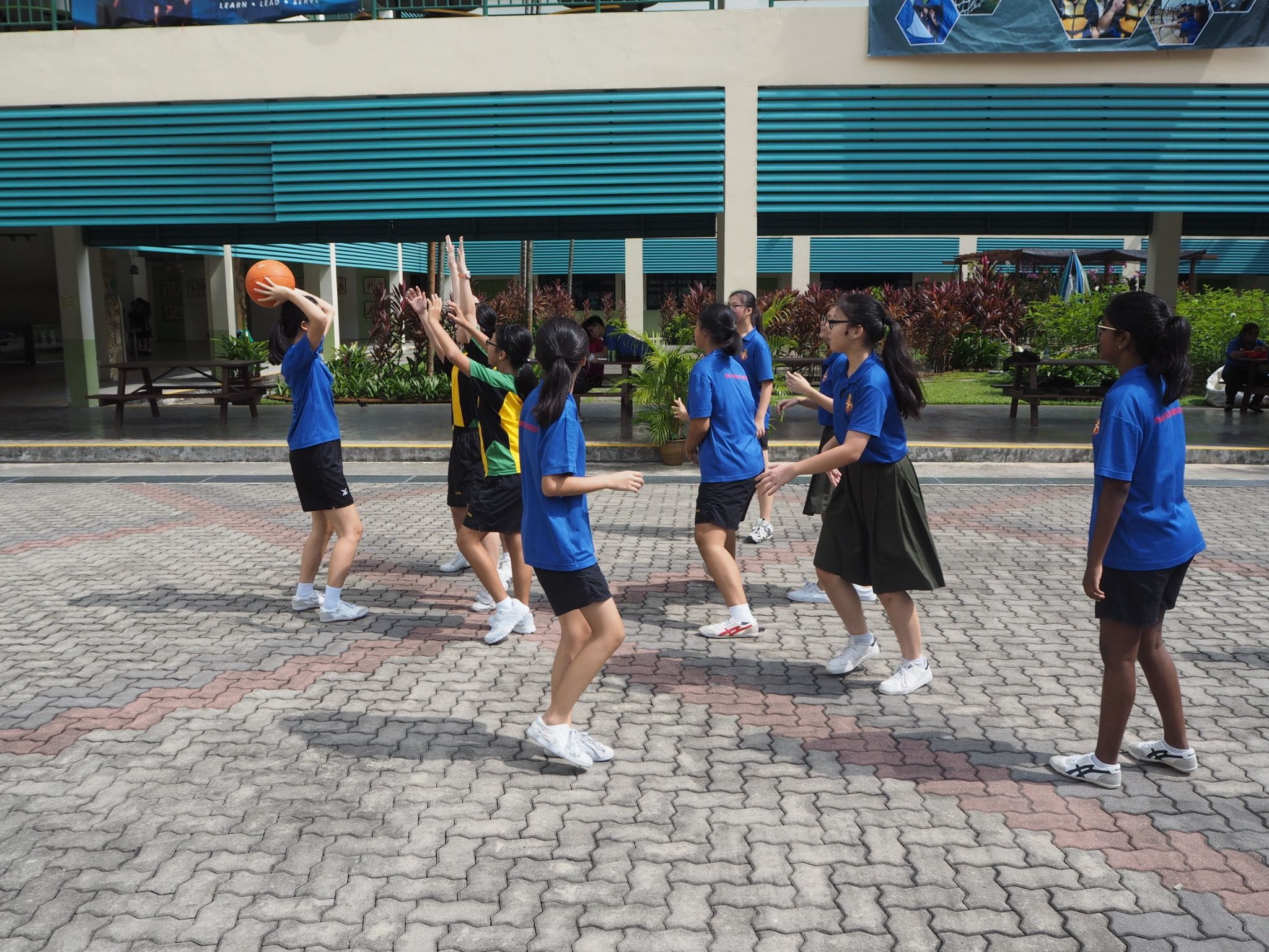 Students in blue uniforms play basketball on a paved outdoor courtyard beside a building with teal louvers.