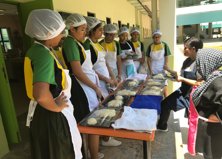 A group of about seven students wearing green and yellow uniform shirts, white hairnets, and aprons stand in a line on an outdoor corridor. They listen attentively to an instructor in a denim apron who is gesturing toward an orange table. Laid out on the table are several black trays holding portions of dough covered in clear plastic wrap.