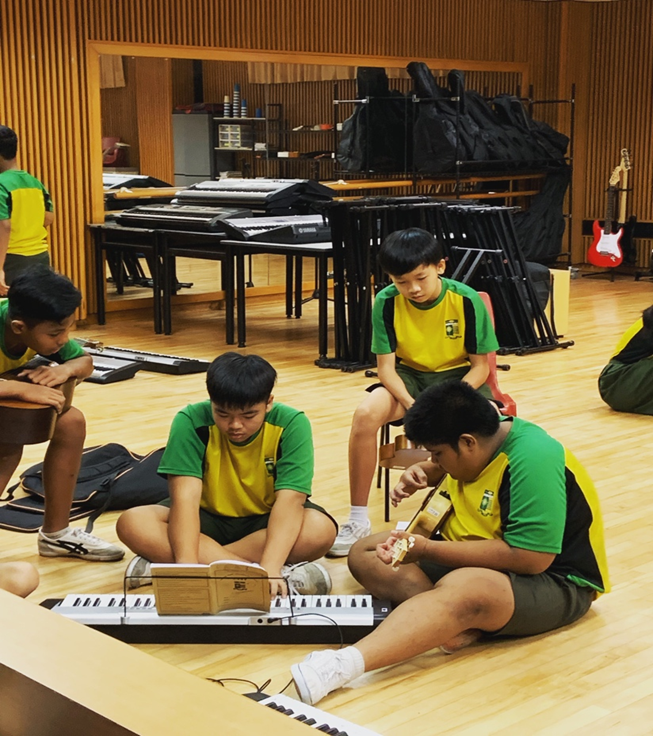 Four schoolboys practice music together on a polished wooden floor in a rehearsal room. Two boys sit cross-legged in the foreground, one reading sheet music while playing a portable electronic keyboard and the other strumming a small guitar or ukulele. In the background, stacks of keyboards, music stands, and instrument cases are neatly arranged near a large wall mirror.