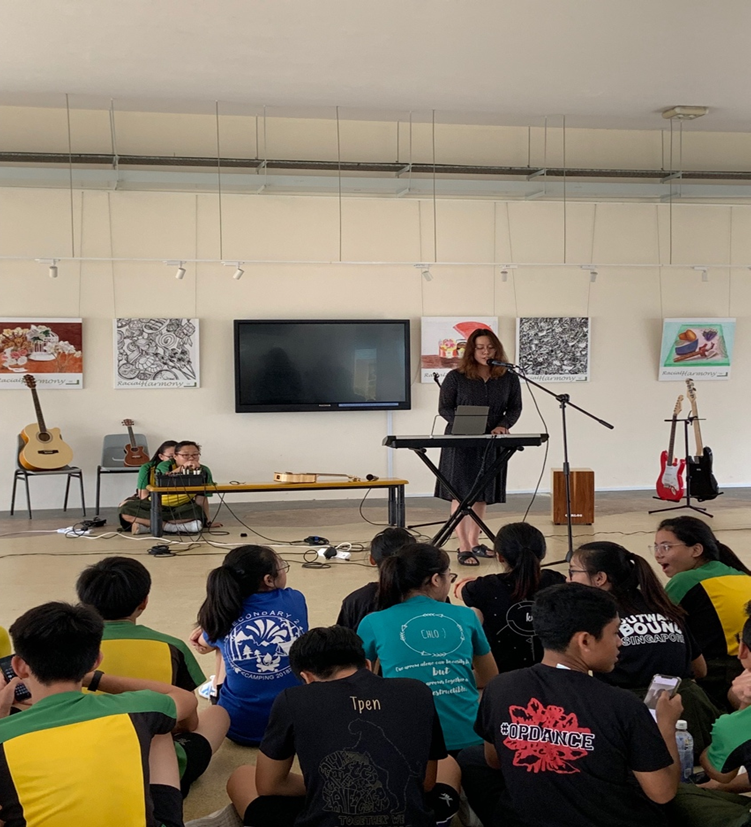 A female instructor stands at a microphone behind an electronic keyboard, presenting to a large group of students sitting on the floor in an open school hall. The wall behind her features a large black TV screen flanked by square art prints reading "Racial Harmony." Acoustic and electric guitars on stands line the sides of the stage area.